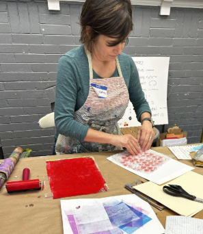 Woman working on fabric project with red paint, paper, and craft supplies on table in a gray room.