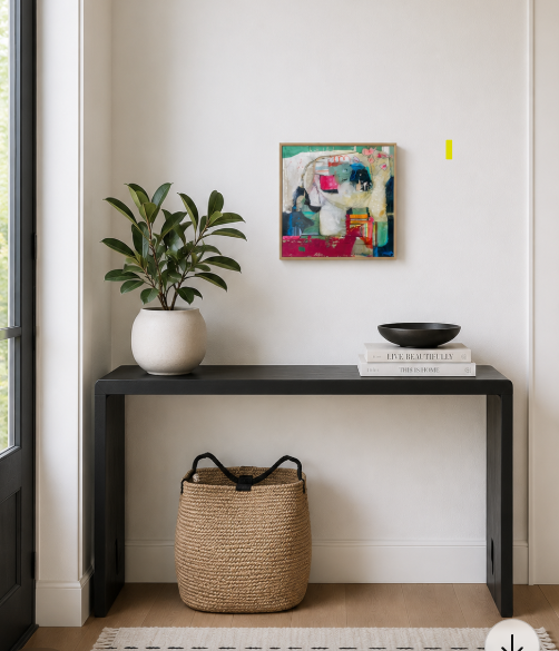 Interior of a room with a black console table against a white wall. On the table, there's a potted plant in a white pot, a black bowl, and two stacked books. A large woven basket sits underneath the table. A colorful abstract painting hangs on the wall above the table. To the left, part of a glass door is visible.