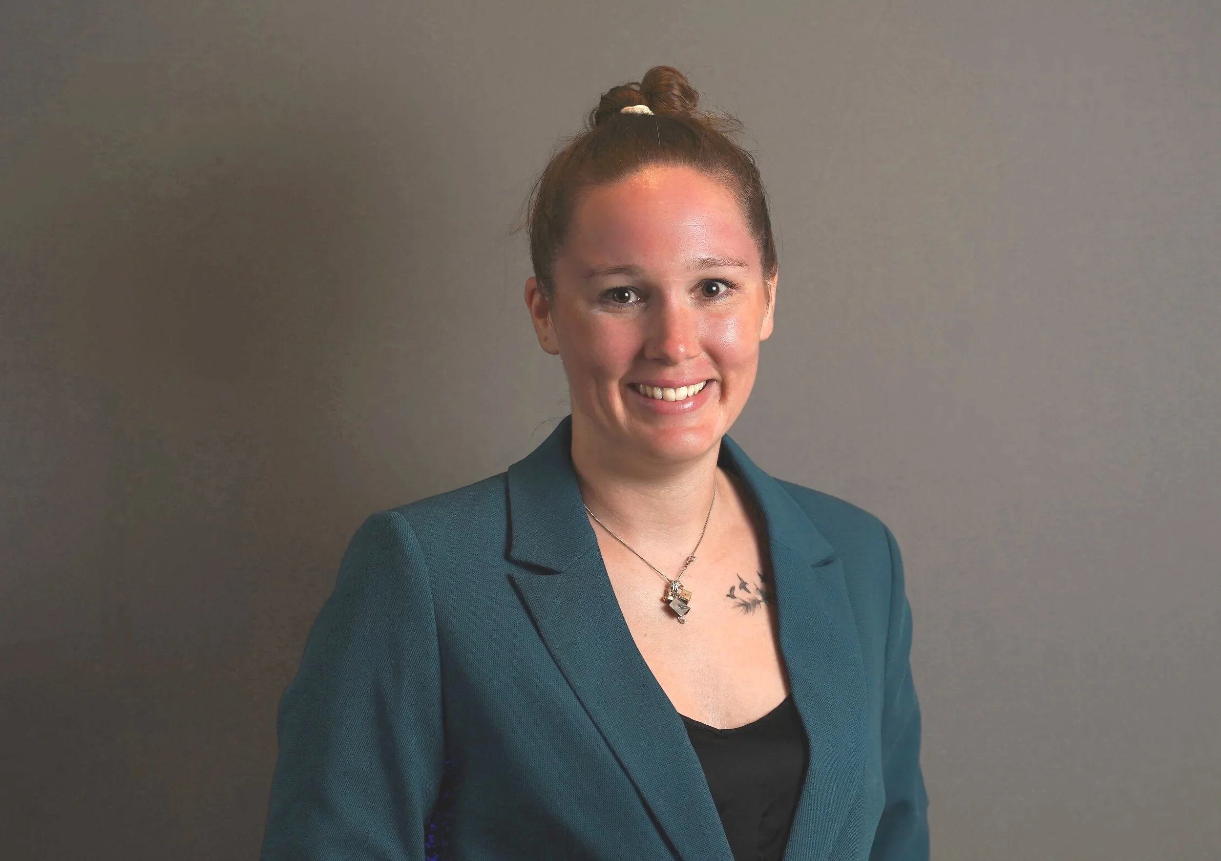 A woman with red hair tied in a bun, wearing a teal blazer over a black top, smiling, with a plain light gray background.