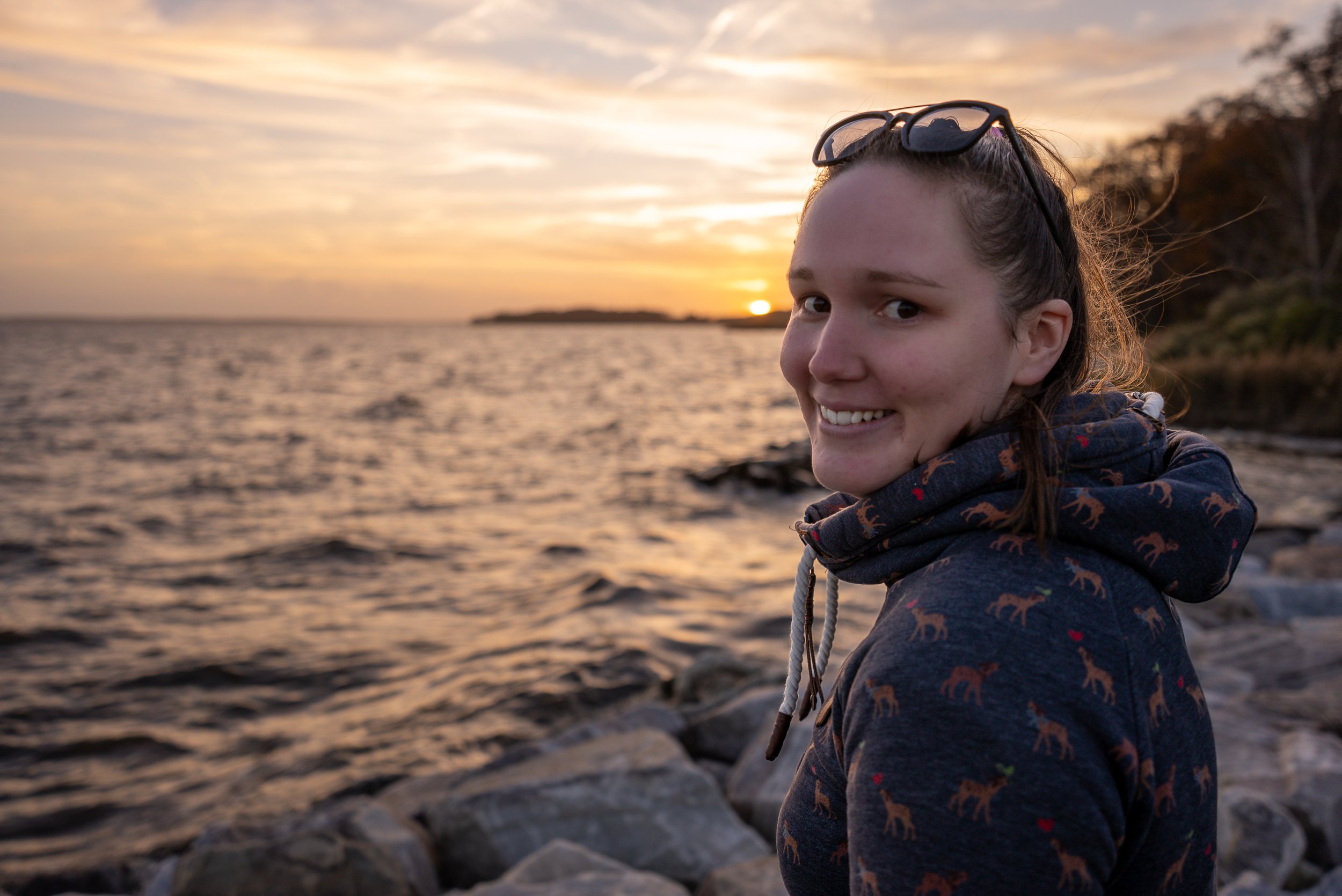 A smiling young girl with sunglasses on her head wearing a jacket with animal print, standing near a rocky shoreline during sunset.