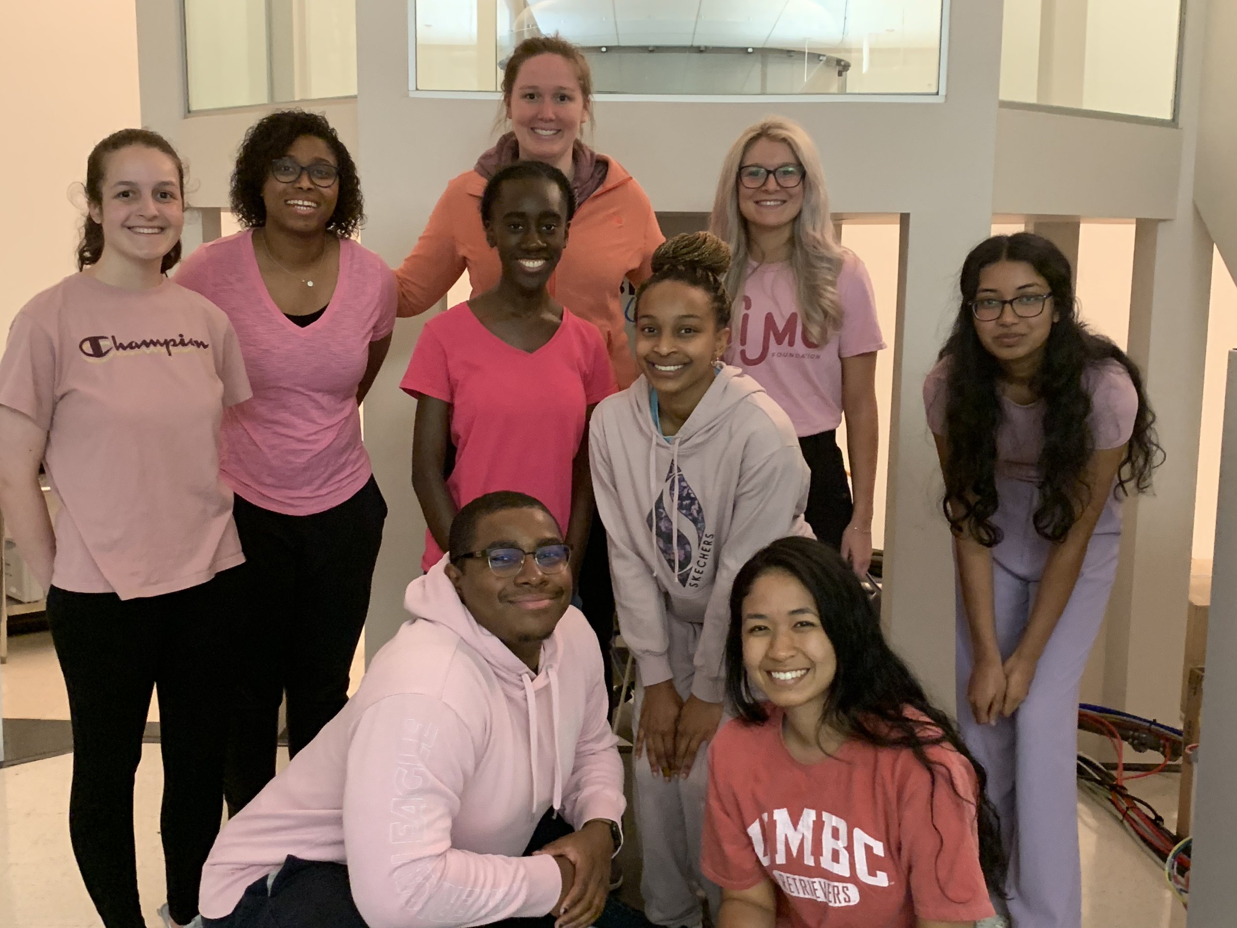 A group of nine researchers smiling and posing together indoors. They are wearing casual clothing, with some wearing pink and red shirts. This is showing a group picture from a scientific research group in front of a NMR magnet.