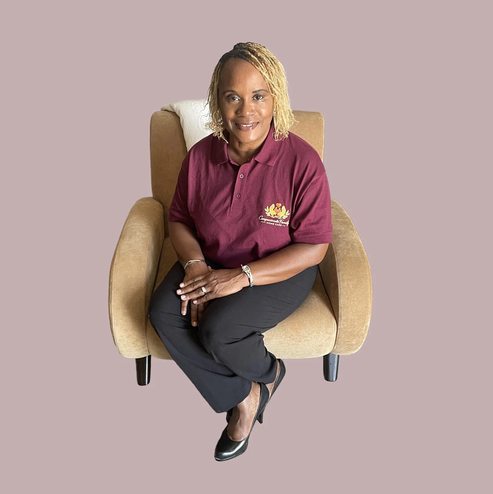 A woman with blonde dreadlocks sitting on a beige armchair with a white pillow, wearing a maroon polo shirt with a logo and black pants, against a light pink background.