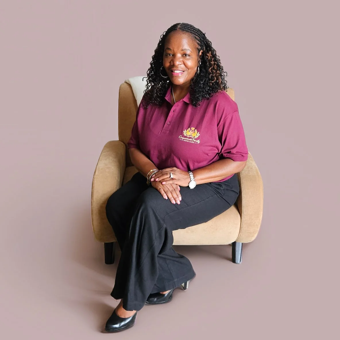 A woman sitting on a beige armchair smiling, wearing a maroon polo shirt with a logo, black pants, and black shoes, against a plain background.