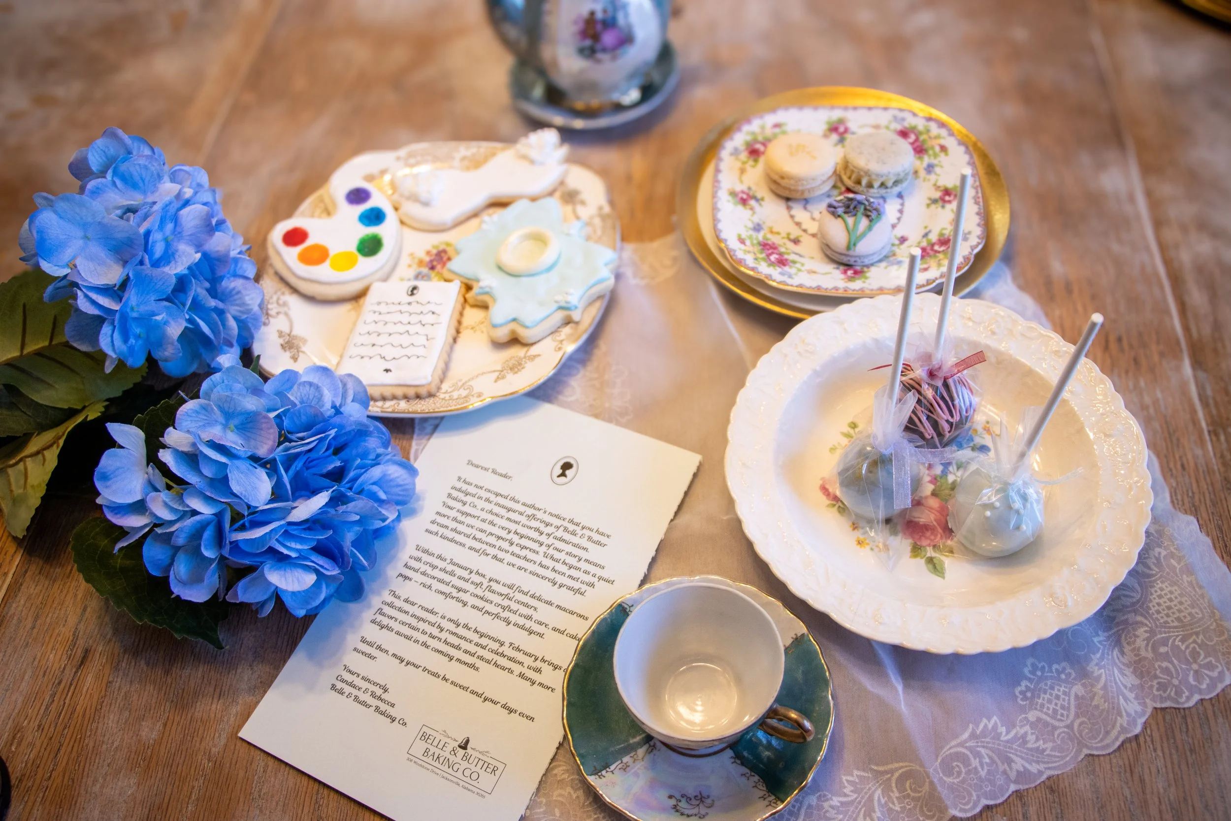An assortment of Belle and Butter Baking Company's treats that were designed for the Bridgerton themed box. There are cookies, macarons, and cake pops in pretty dishes, as well as tea cups and a teapot, and a Lady Whistledown letter.