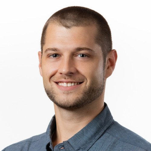 A young man with short brown hair, light beard, wearing a blue button-up shirt, smiling against a plain white background.