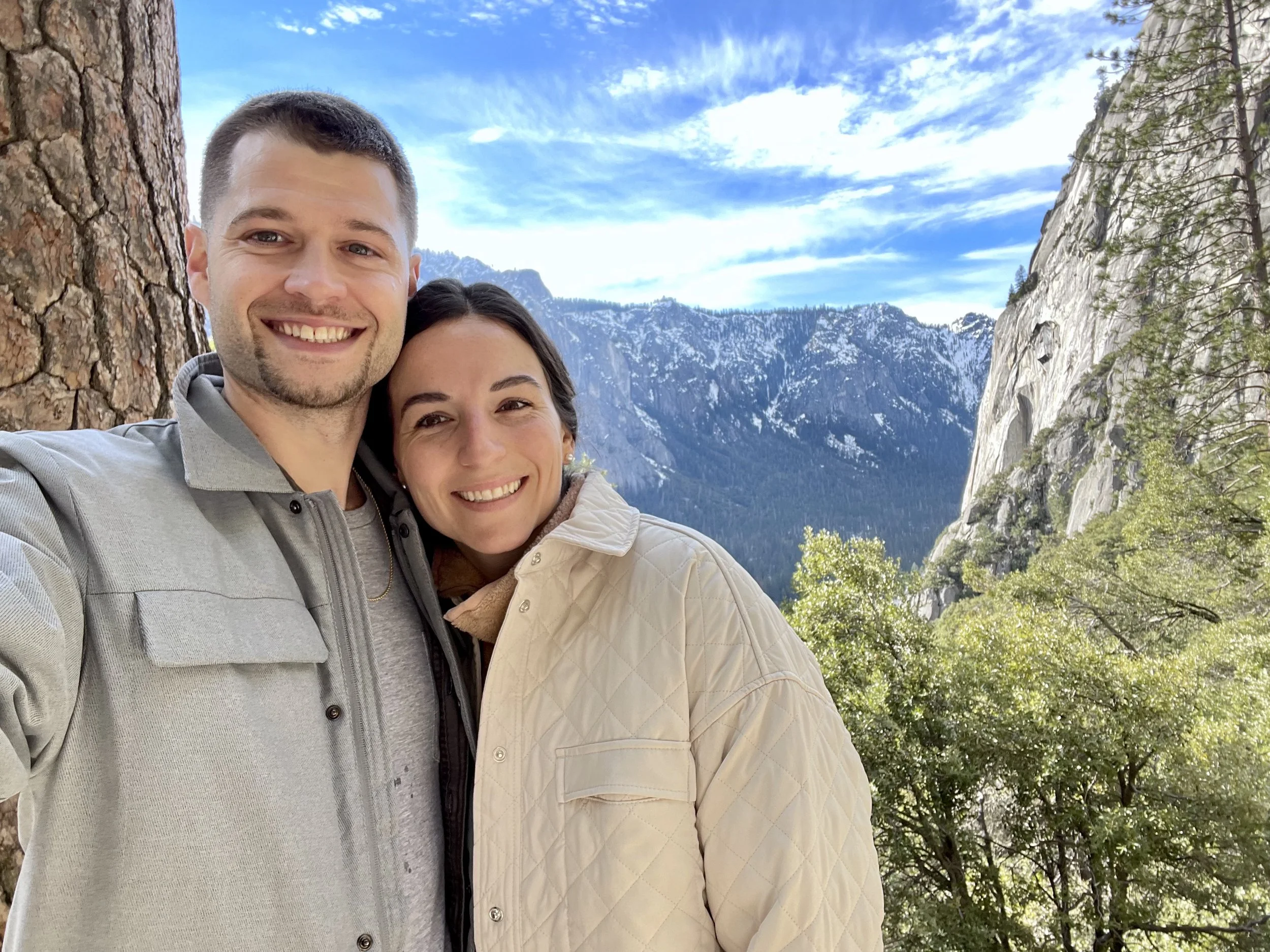 A smiling man and woman taking a selfie outdoors with mountains and a blue sky in the background.