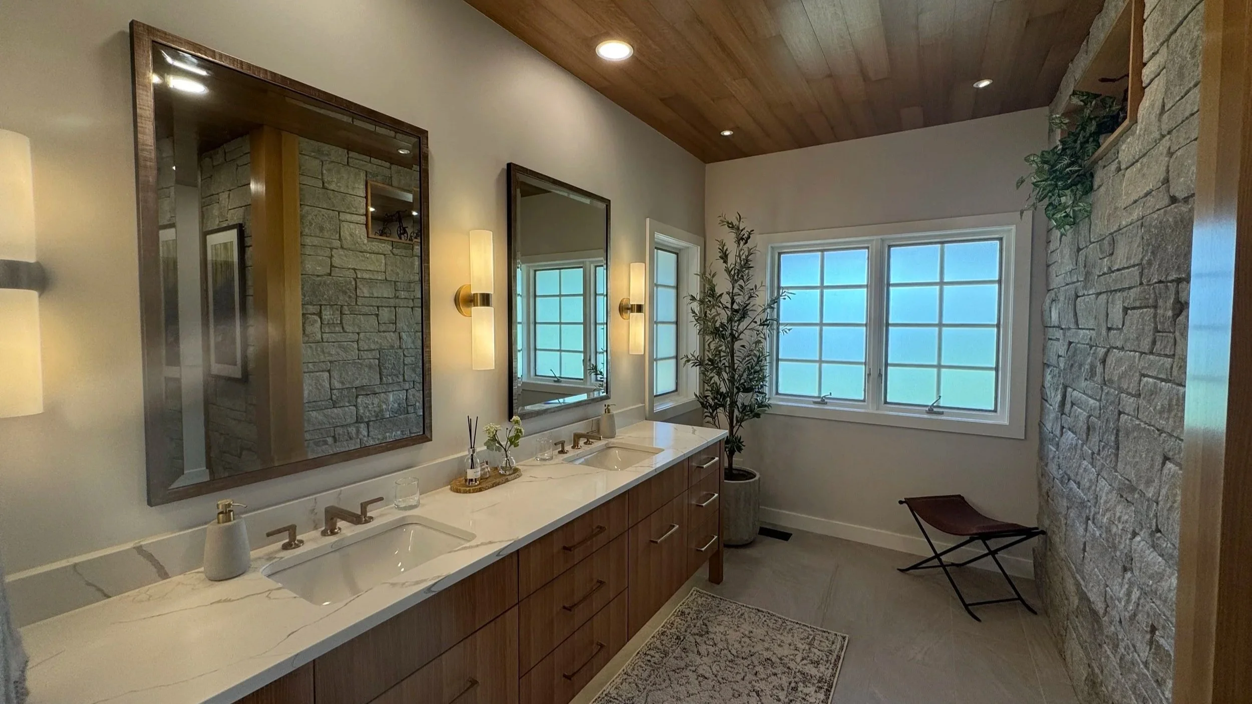 Modern bathroom with double vanity, large mirror, stone accent wall, large windows, and wooden ceiling.