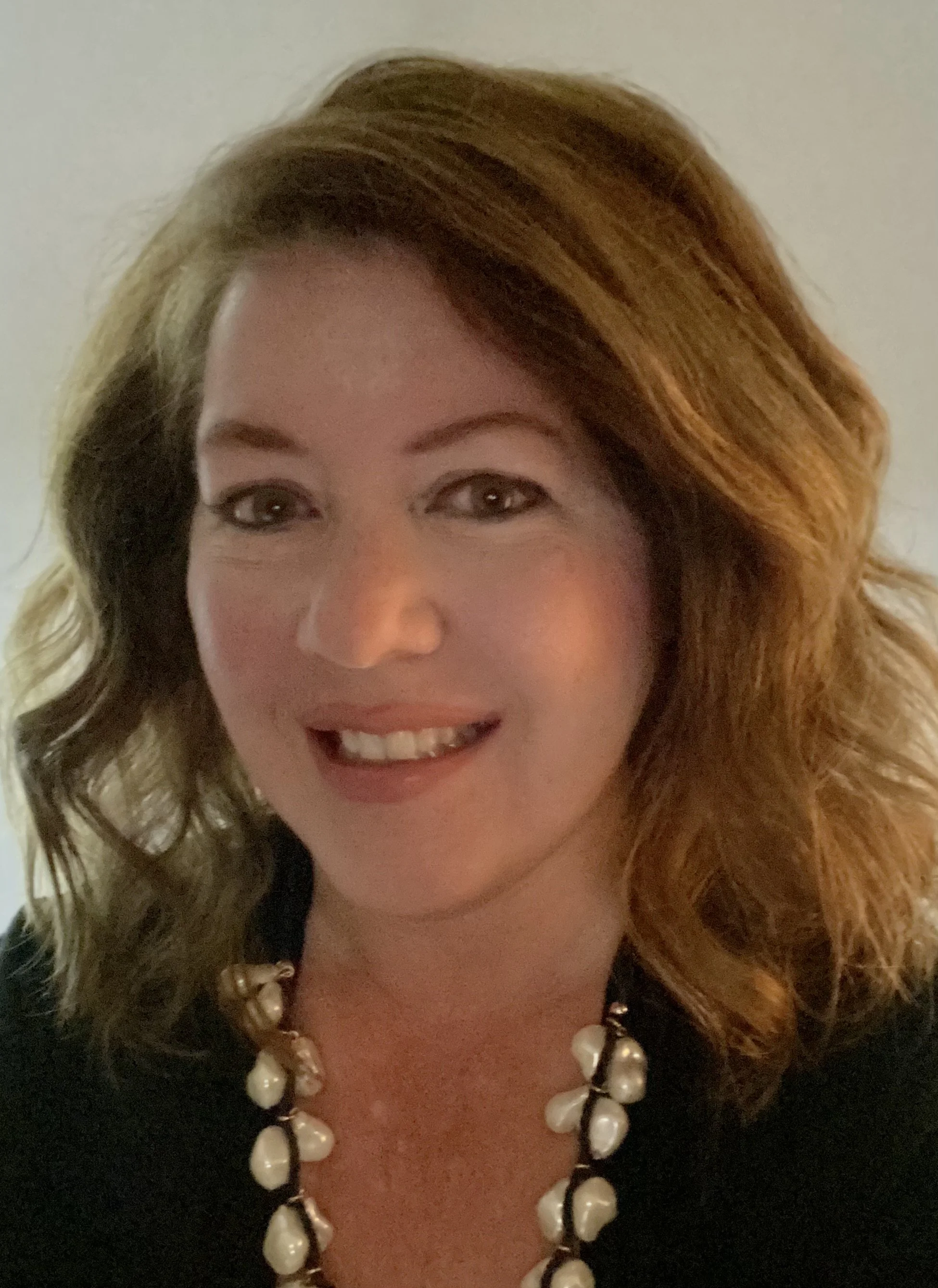 A woman with long, wavy hair, smiling, wearing a pearl necklace and a black top.