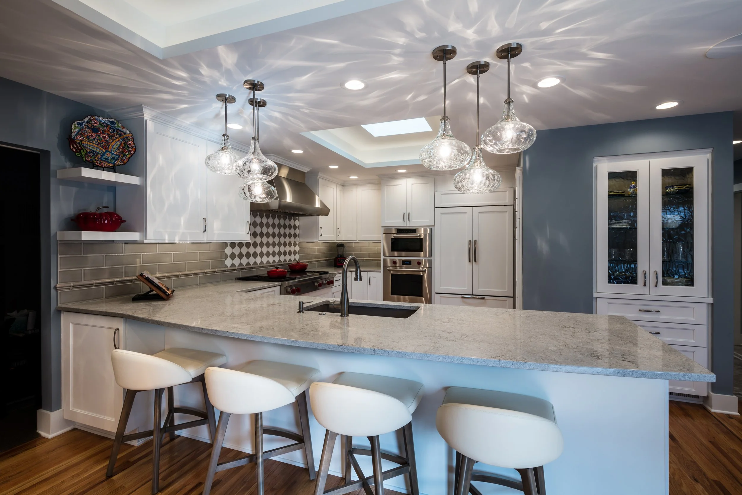 Kitchen with white cabinets, stainless steel appliances, beige tile backsplash, granite island with white chairs, pendant lights, and a skylight.