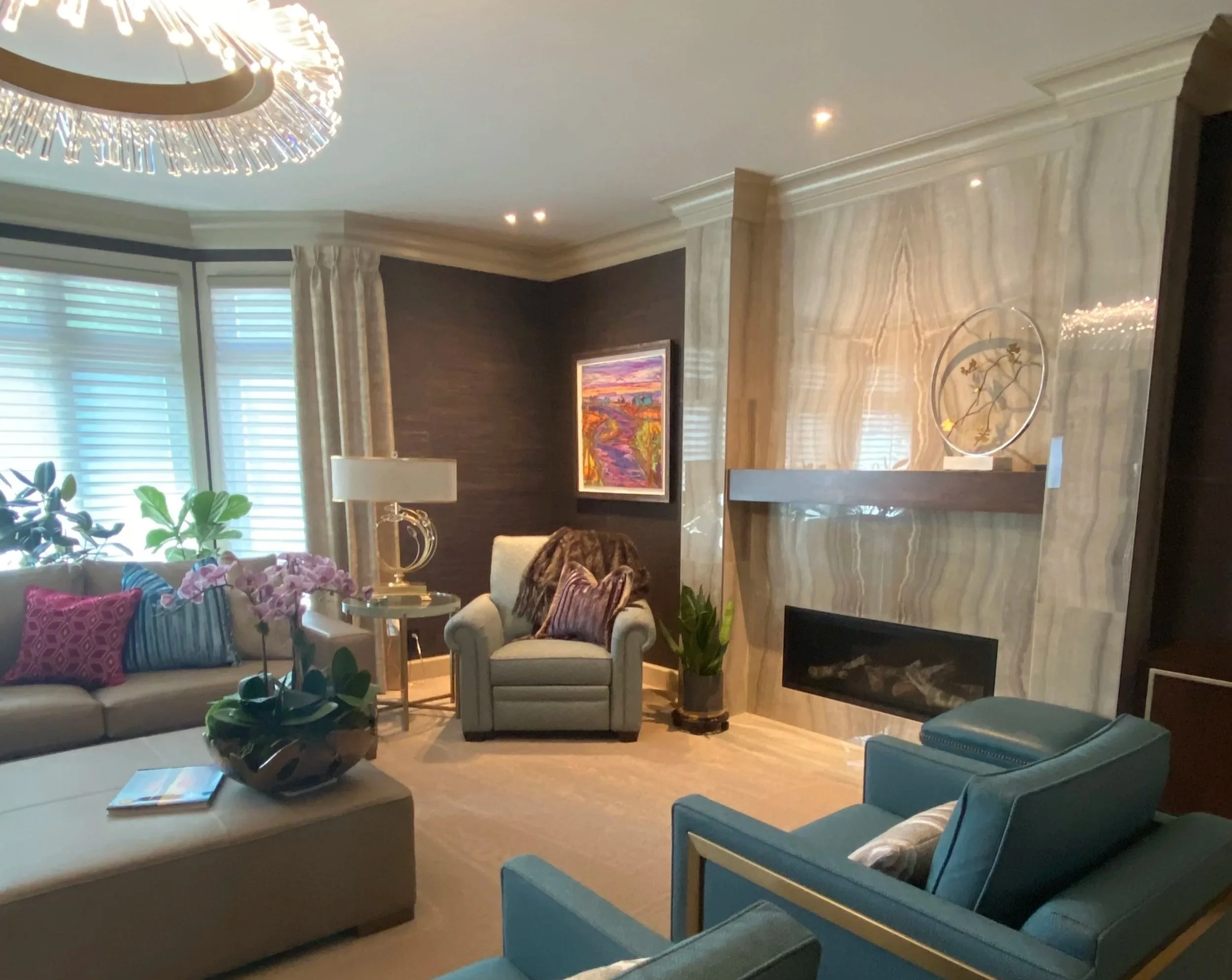 A living room with a white sofa and armchair, colorful throw pillows, a coffee table with a plant and magazine, a marble fireplace, framed artwork on the dark-colored wall, large windows with blinds and curtains, and a chandelier on the ceiling.