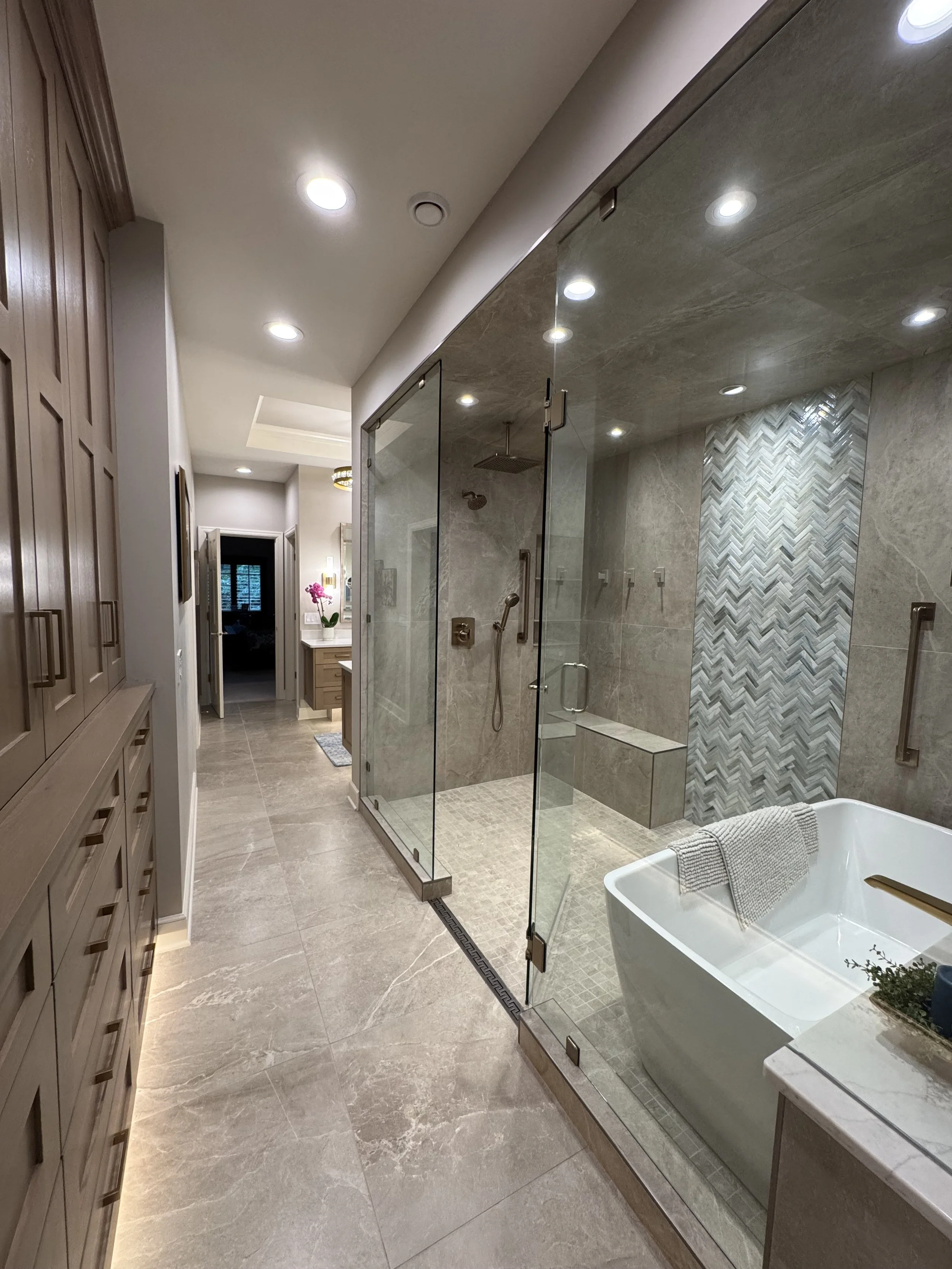 A modern bathroom with a glass-enclosed shower, a freestanding bathtub, wooden cabinetry, and beige marble-like tiles on the floor and walls, illuminated by ceiling lights.
