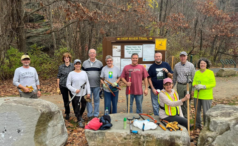 Group of ten people standing and sitting outdoors on a trail, holding tools and equipment, posing for a photo in front of a trail sign in a wooded area during autumn.