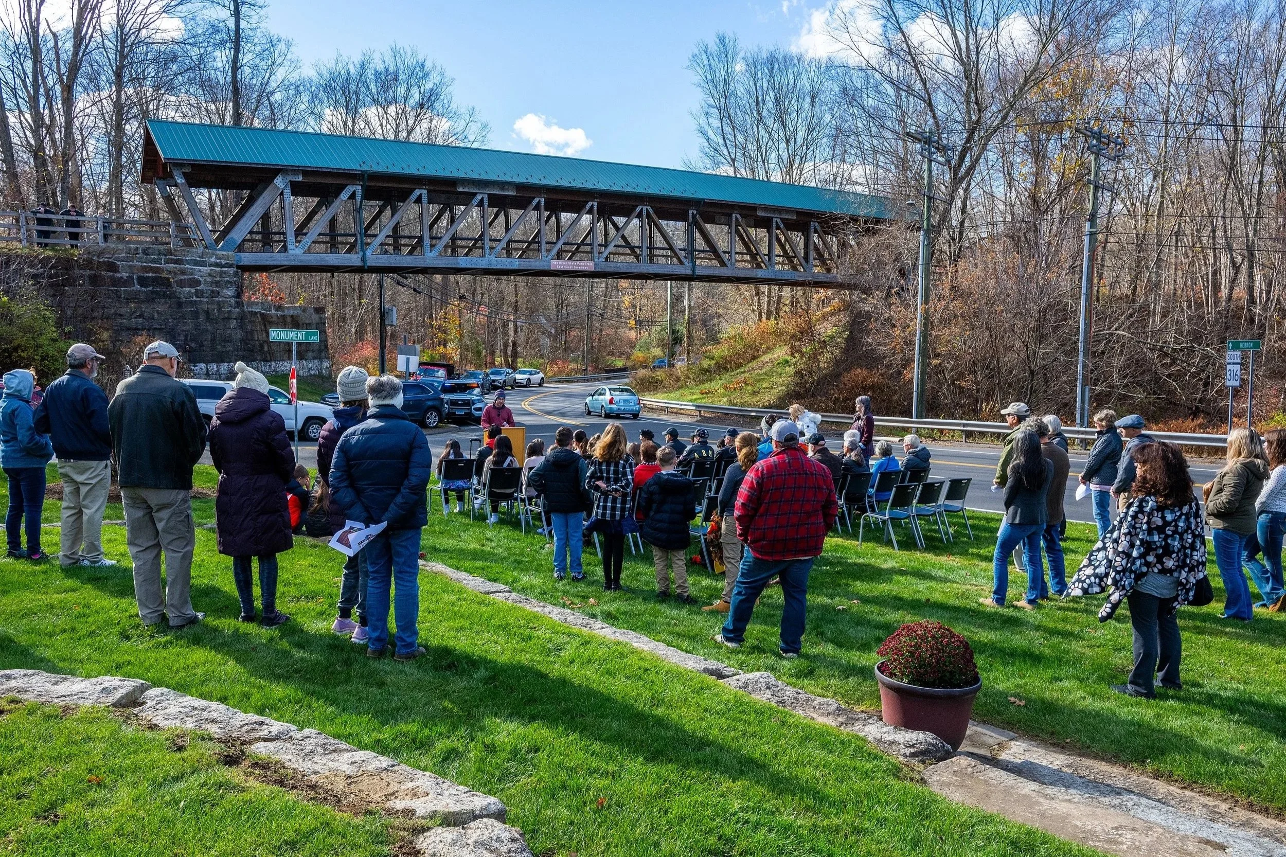 Gathering of people outdoors near a blue metal covered bridge, with some people seated and others standing, during daytime with clear skies and leafless trees in the background.