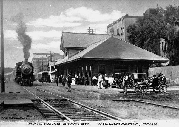 Historical black-and-white photo of the Wilmington, Connecticut railroad station with a steam train arriving and people standing outside.