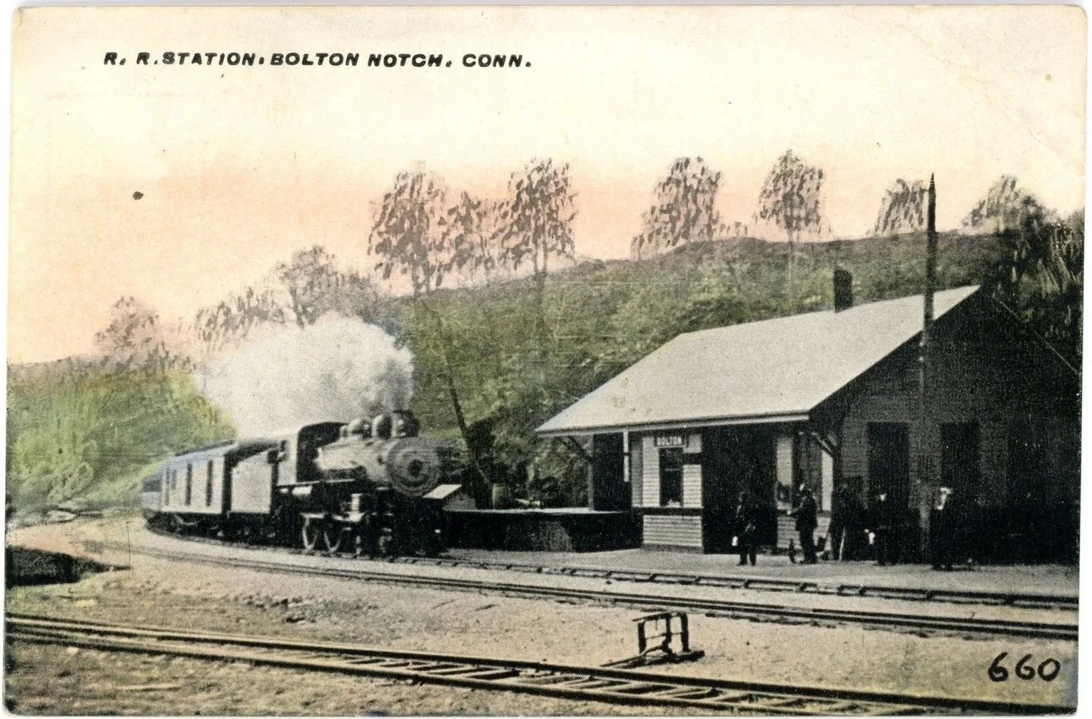 A vintage black and white photograph of a steam train approaching a small station platform with a building and a few people standing nearby, set against a backdrop of trees on a hillside.