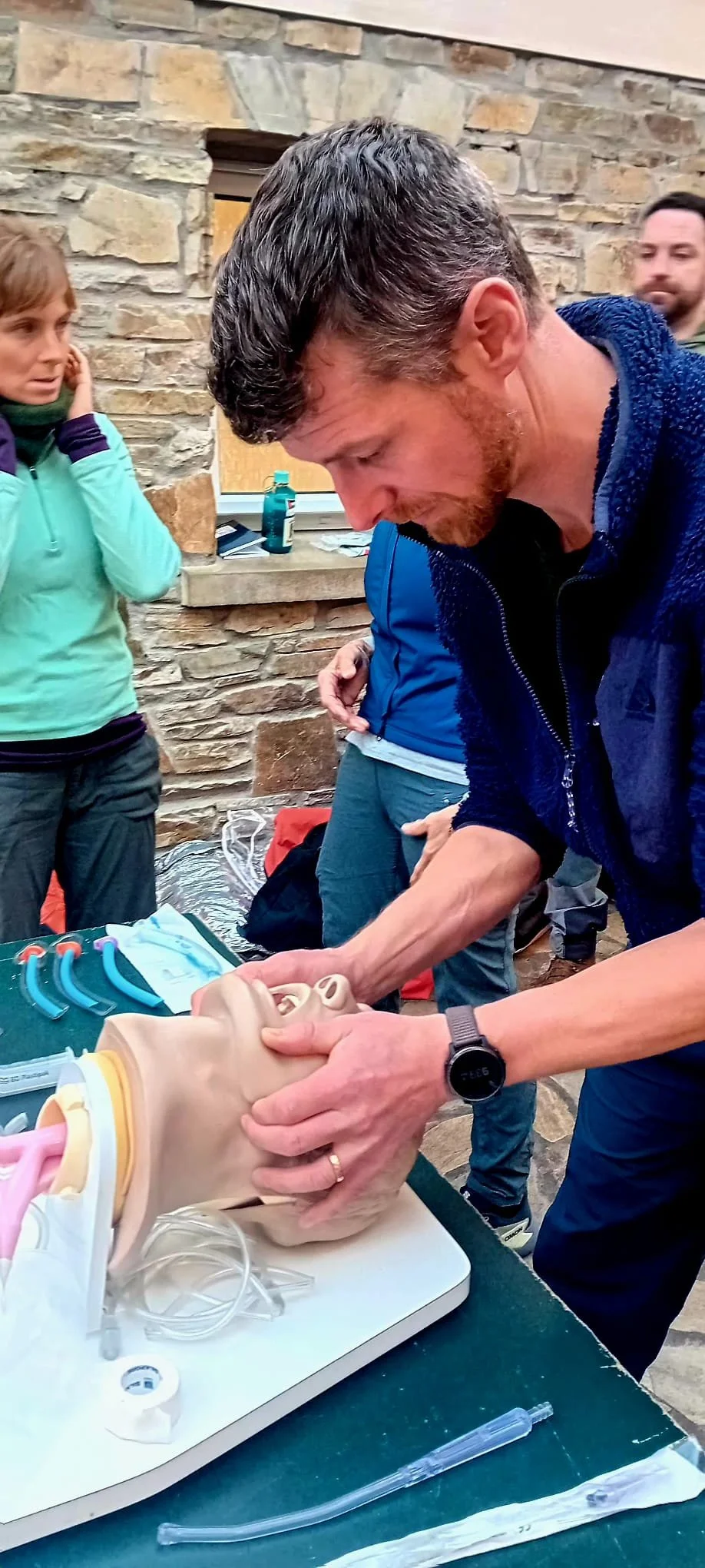 A man practicing CPR on a medical training dummy during a training session, with onlookers watching.