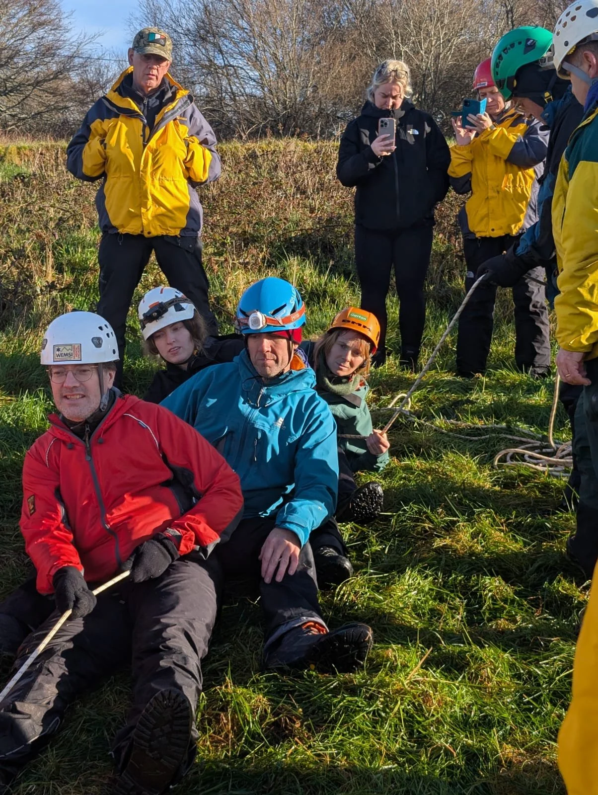 Rescue team members in outdoor setting, some sitting on the grass in climbing gear, others standing, taking photos, and observing.