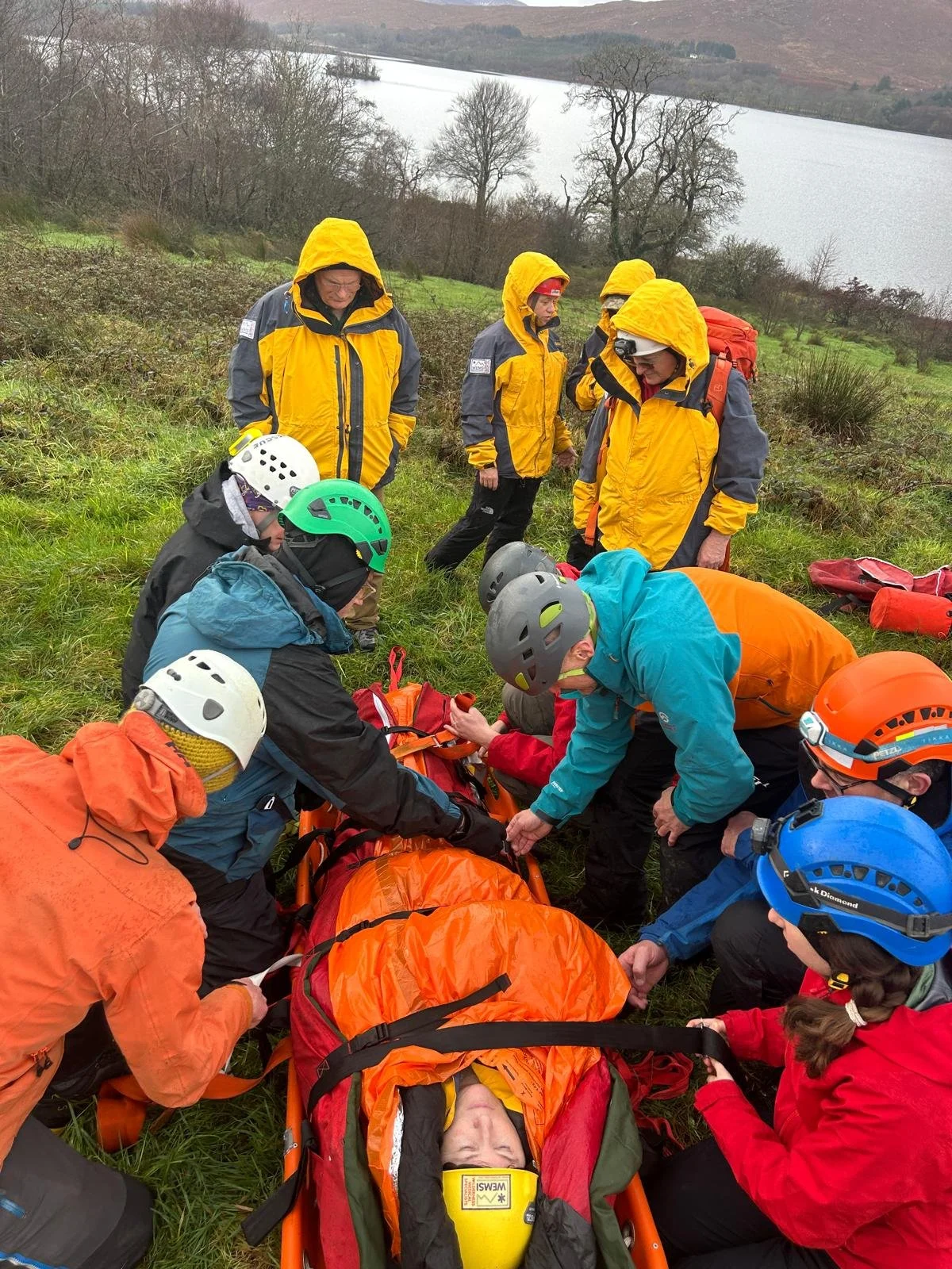 Group of rescue team members attending to a person lying on a stretcher in a grassy outdoor area near a lake, wearing helmets and outdoor gear.