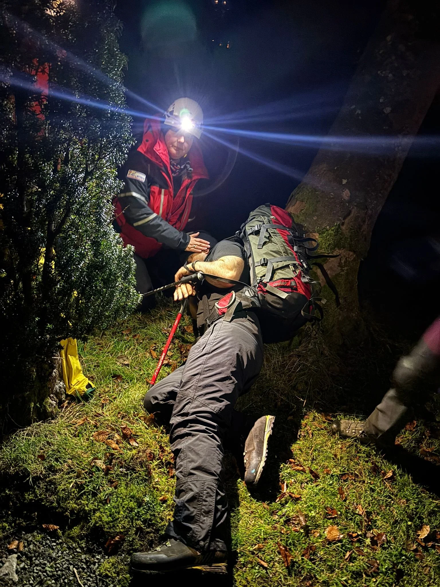 Nighttime rescue scene with a person lying on the ground, surrounded by trees and grass, with another person, wearing a headlamp, attending to them. The rescuer has a headlamp, red and black jacket, and gray pants with a backpack, indicating they are
