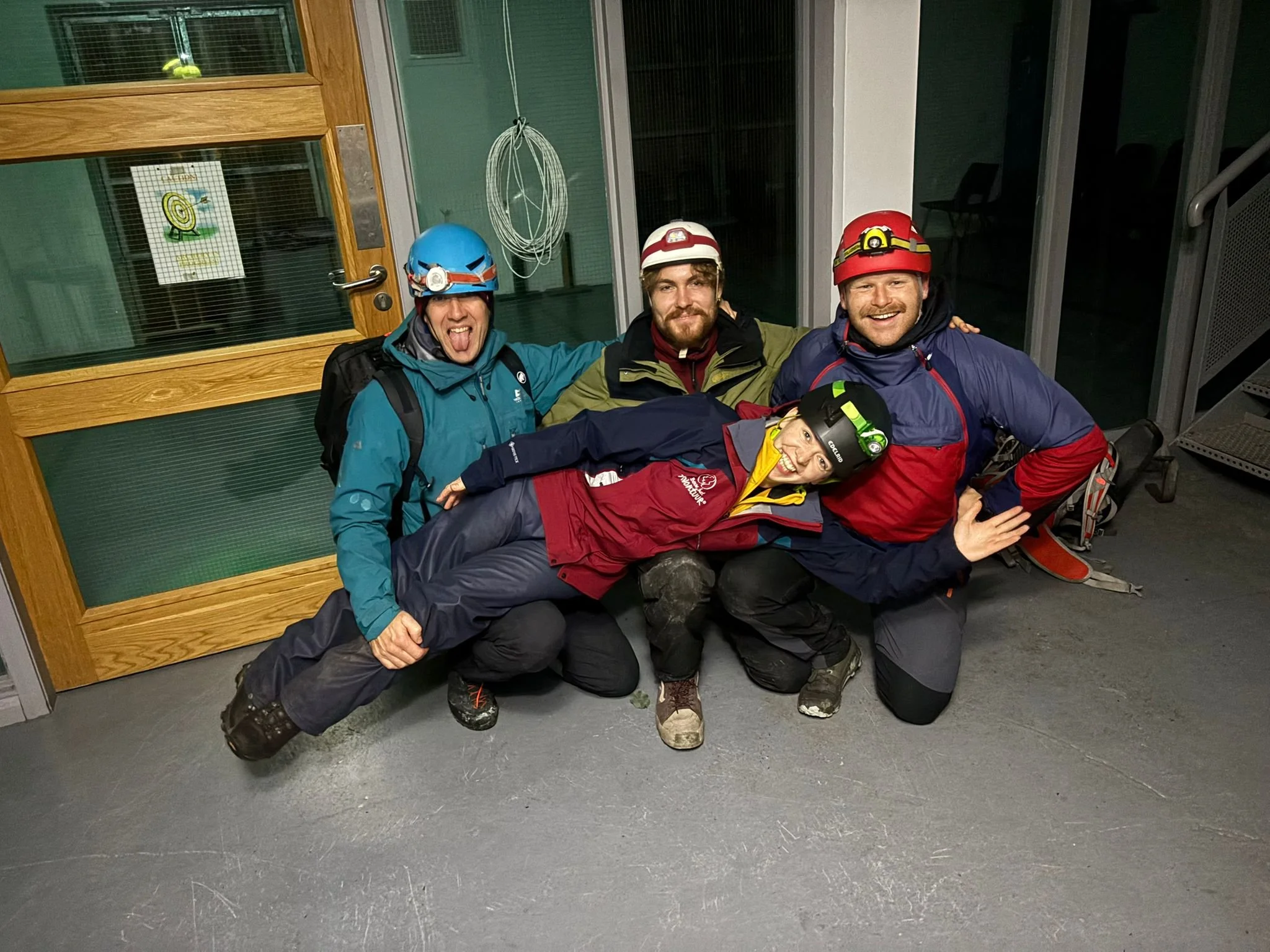 Four people dressed in outdoor gear and helmets, smiling and posing together indoors with a glass door in the background.