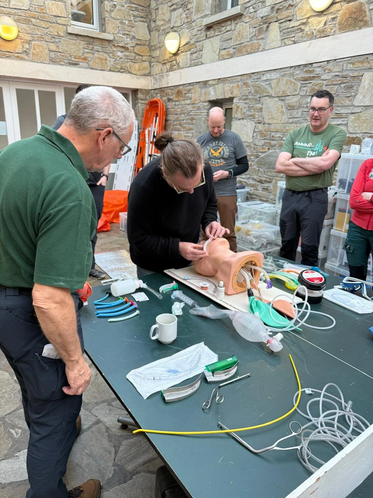 Group of people observing a medical training session with a man practicing intubation on a medical mannequin on a table filled with medical instruments and supplies.