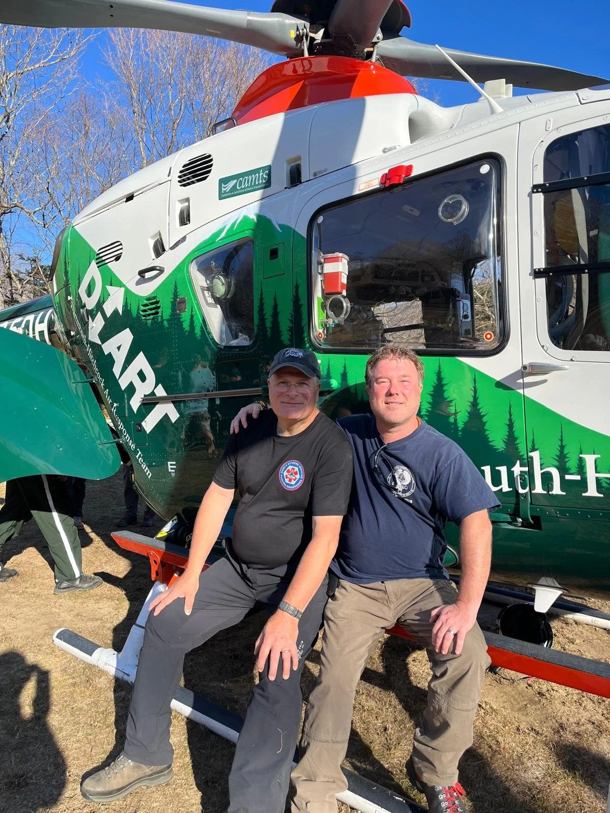 Two men sitting on a helicopter landing skids in front of a rescue helicopter, with trees and a clear blue sky in the background.