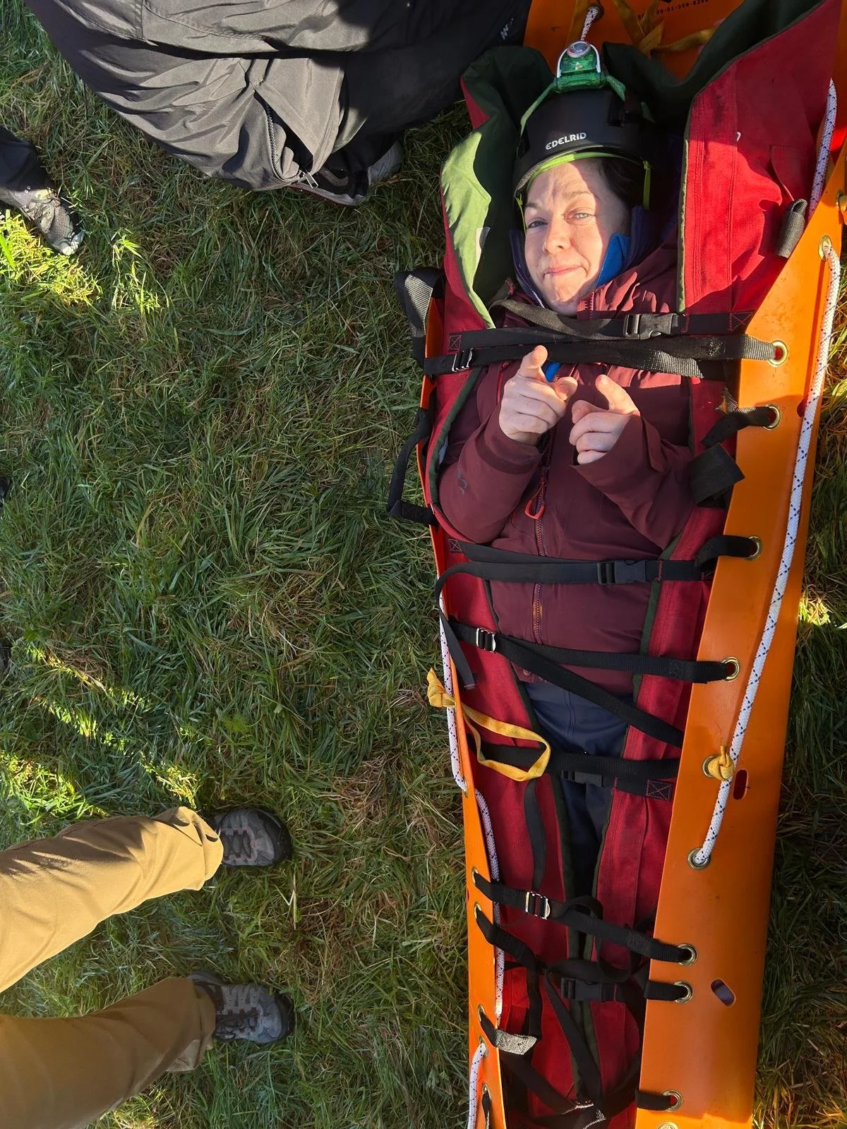 Person lying on an orange rescue sled on grass, giving a thumbs-up and pointing with both hands, wearing a headlamp and outdoor gear.