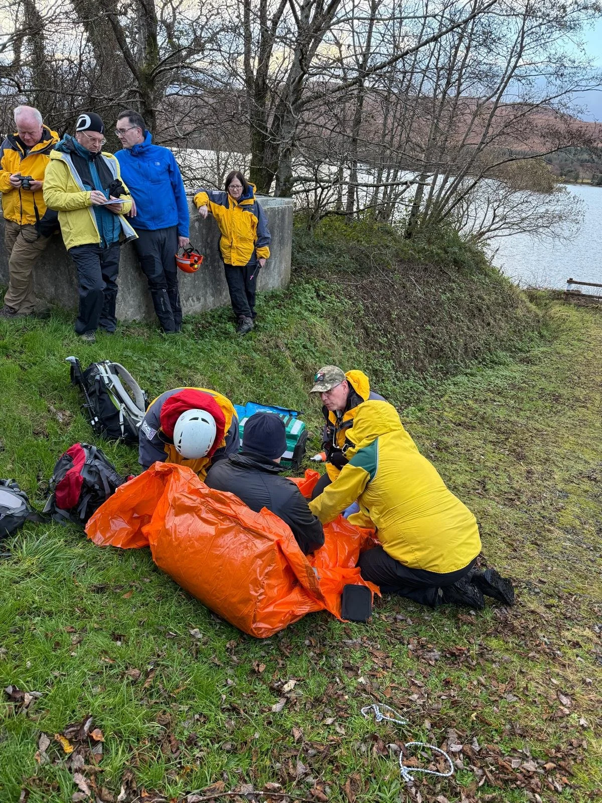 Group of people in yellow jackets assisting a person with a rescue or medical emergency outdoors near a lake, with trees and cloudy sky in the background.