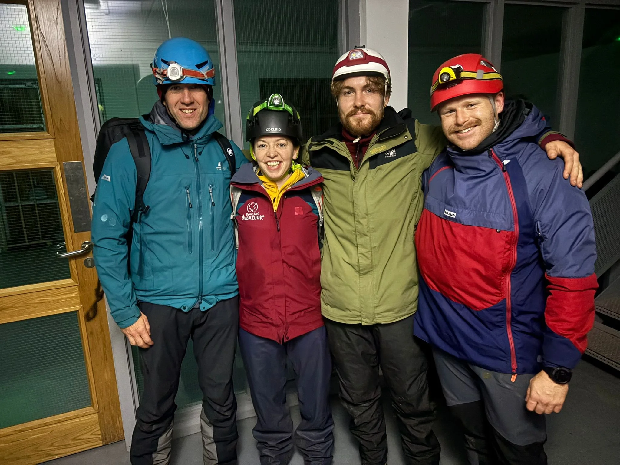Group of four people in outdoor gear, wearing helmets and harnesses, standing indoors in front of a glass window.