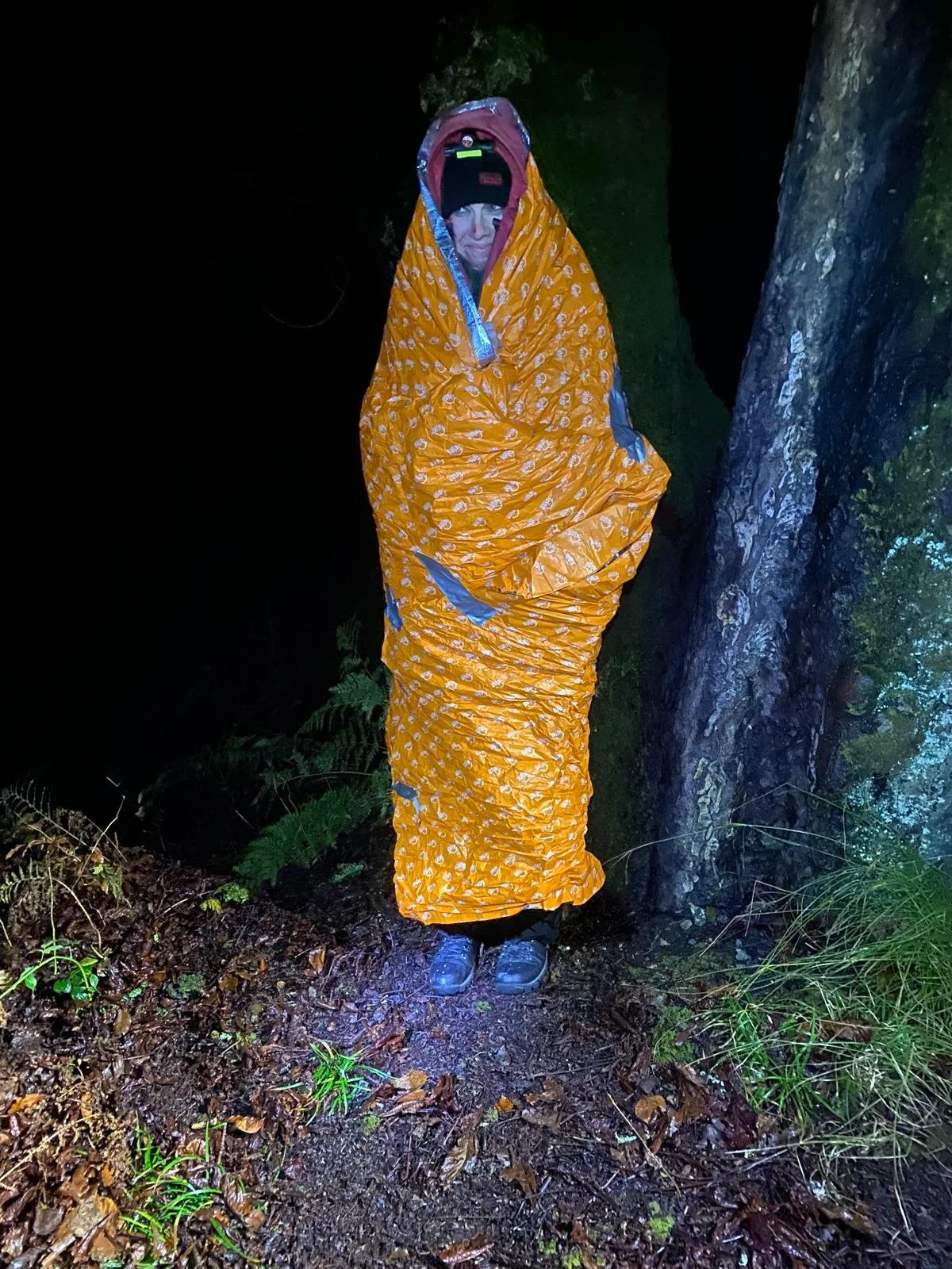 Person standing outdoors at night, wrapped in an orange sleeping bag with a shiny pattern, wearing a black hat and sturdy shoes, near a tree and green foliage.