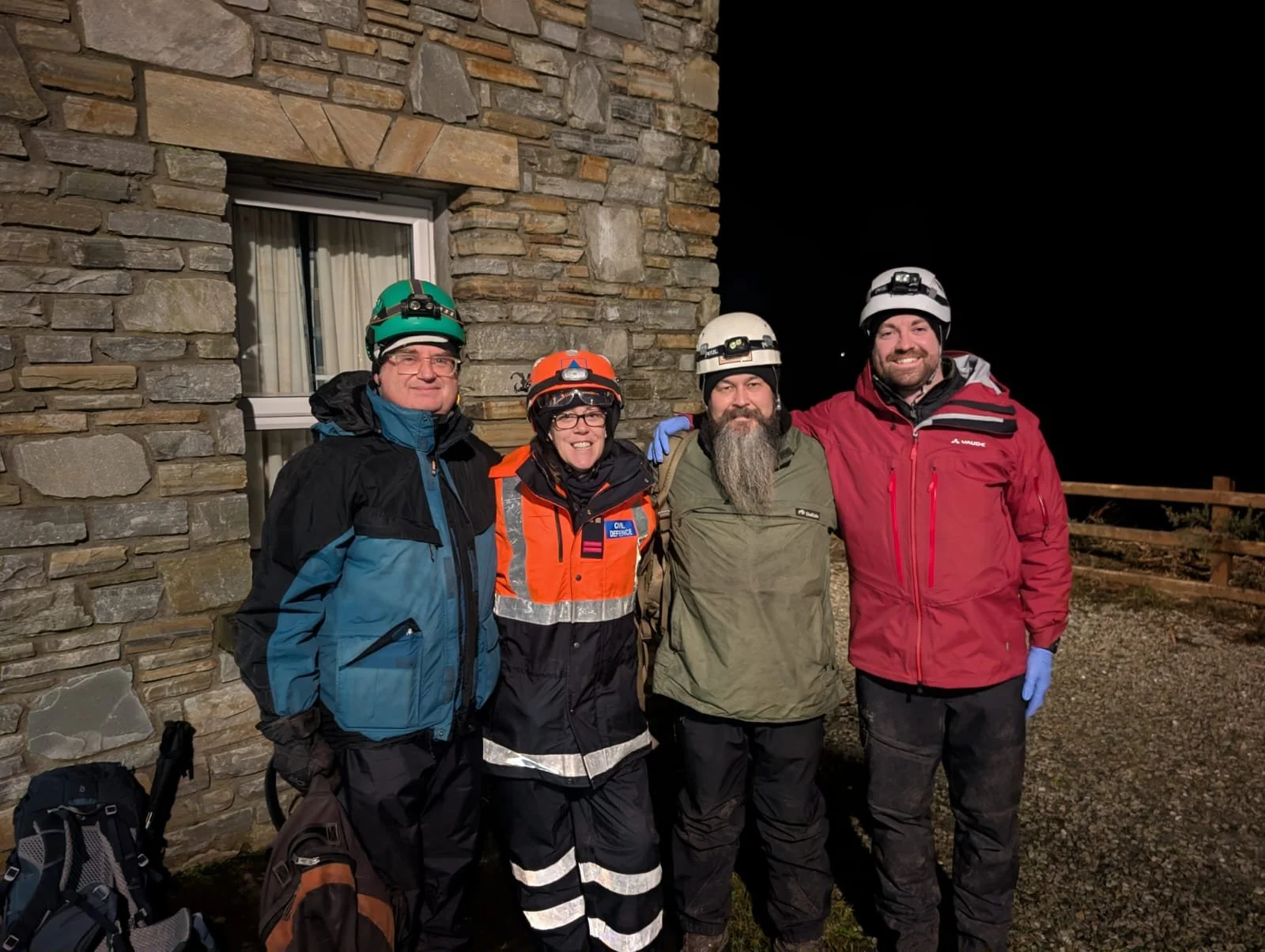 Four people in protective gear and helmets standing outdoors at night against a stone building wall, smiling for the photo.