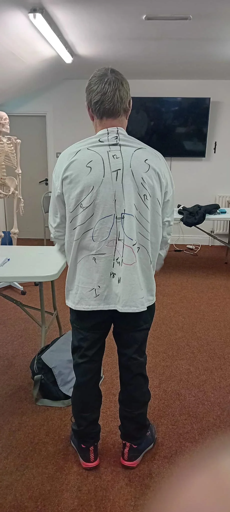 Person standing in a room with medical diagrams of the human body drawn on their back. Skeleton visible to the side. Room has tables, a television, and teaching equipment.