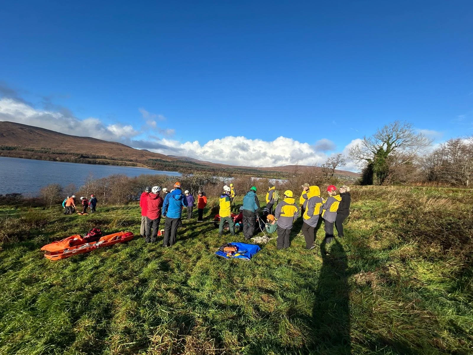 Group of outdoor enthusiasts preparing for water rescue training near a lake, with equipment on the grass, under a partly cloudy sky.