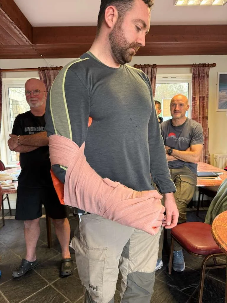 A man with a beard and wearing a dark long-sleeve shirt stands inside a room. His left arm is wrapped in pink bandages, and he looks down at it. Two other men are in the background, one with glasses and a black T-shirt, and the other with a gray T-sh
