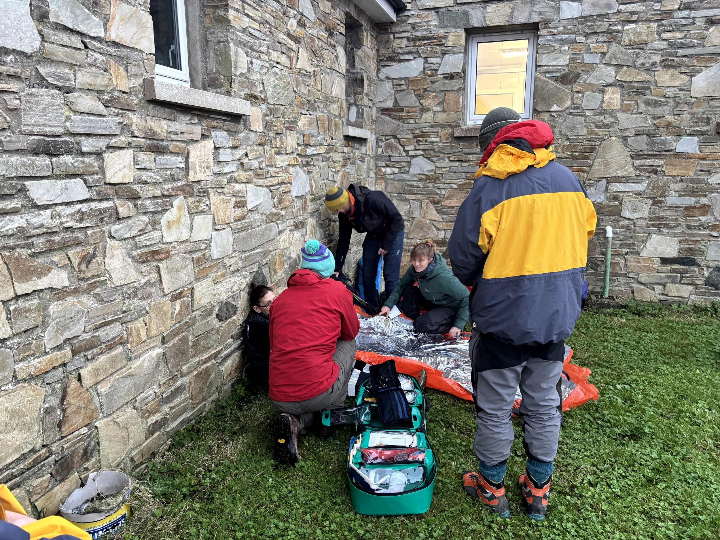 Group of people setting up a tent outside a stone building on a grassy area, with tools and equipment visible.