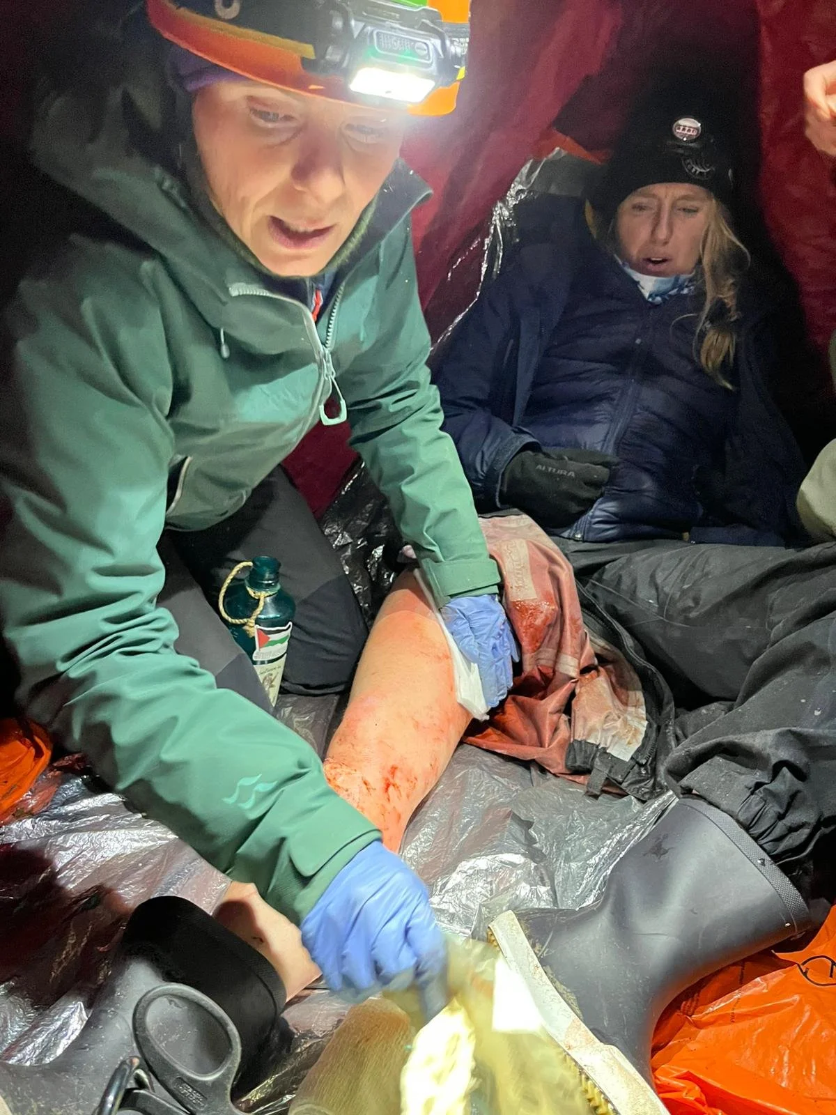 Rescue workers providing first aid to an injured person with a bloodied leg inside a tent or shelter in a mountainous area, wearing headlamps and outdoor gear.
