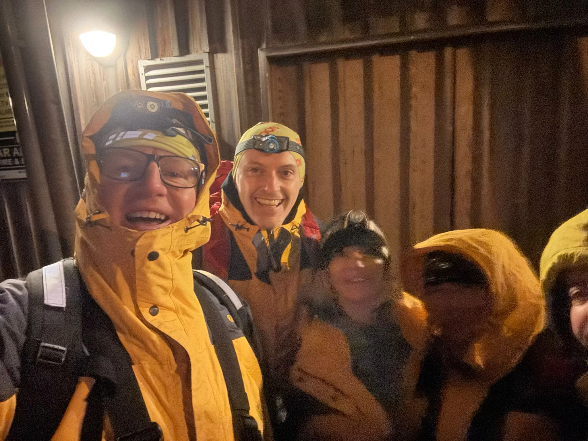 Four people dressed in outdoor gear with yellow jackets and headlamps, smiling inside a wooden structure at night or in a dimly lit area.