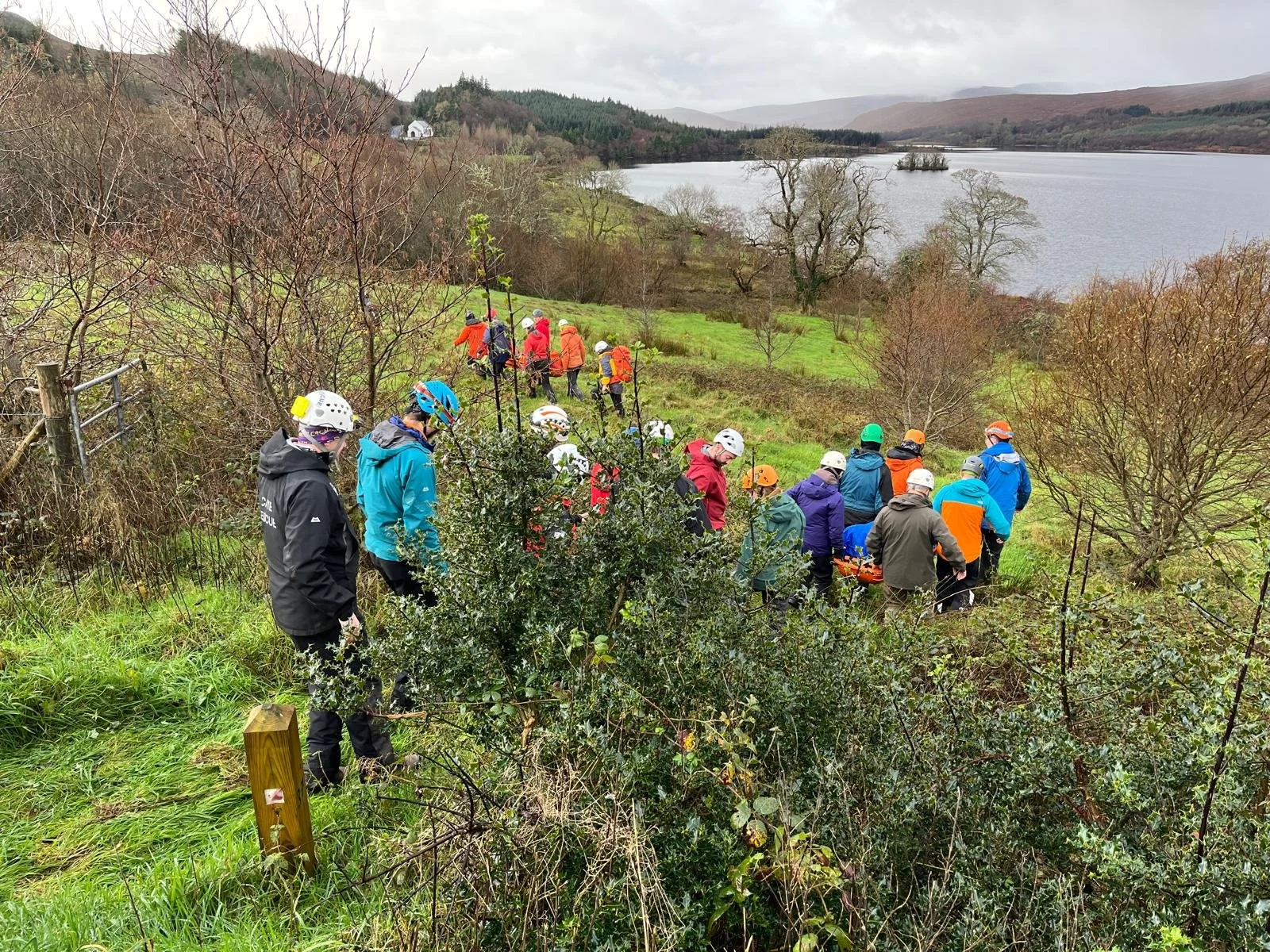 A group of people wearing helmets and outdoor clothing walking down a grassy, bushy hillside near a lake with trees and mountains in the background.