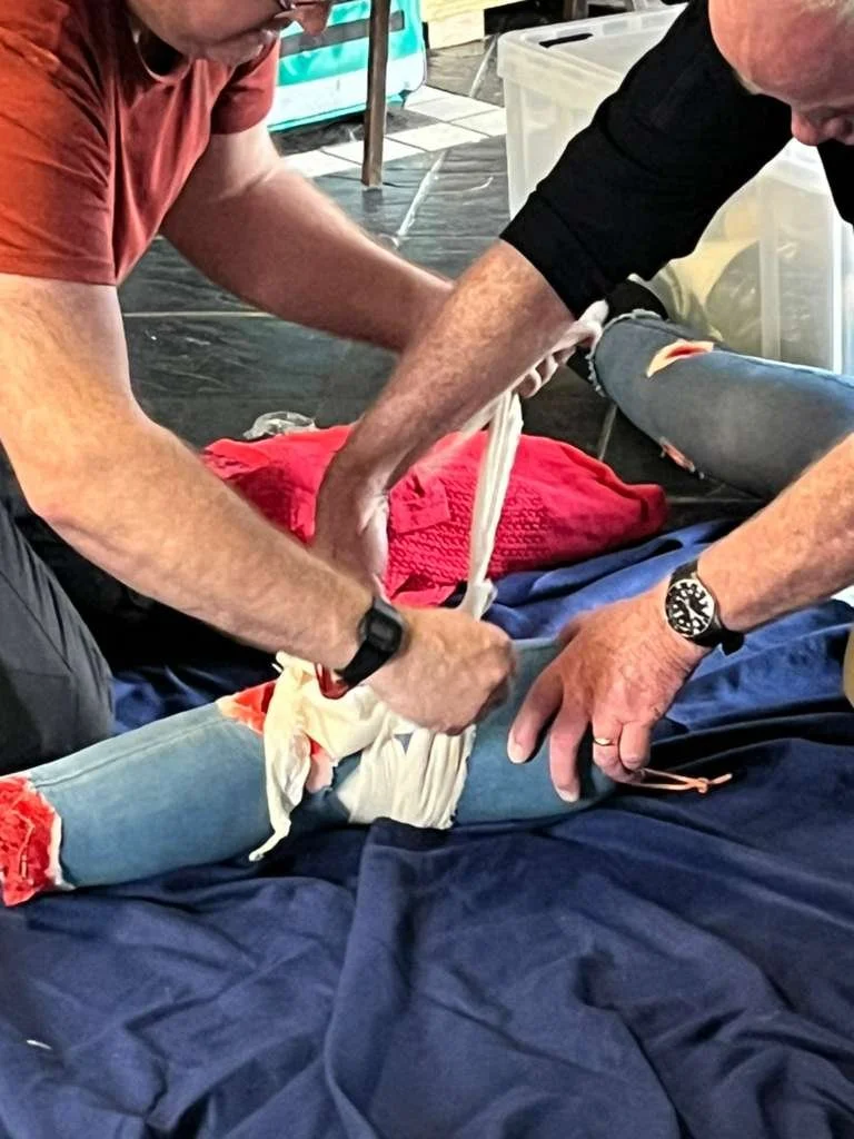 Two men assisting a person who appears to be injured or in distress, with the man on the left holding a makeshift sling for the person's leg.