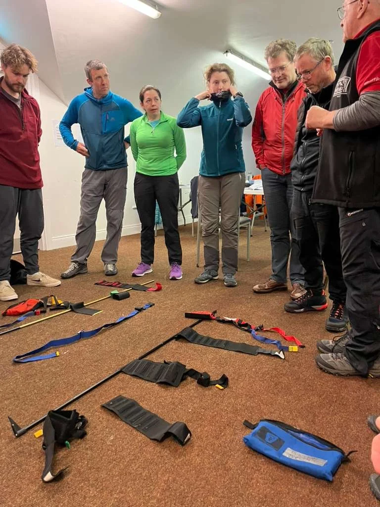 Group of seven people standing in a circle, observing search and rescue equipment laid out on the floor, including harnesses, poles, and straps.