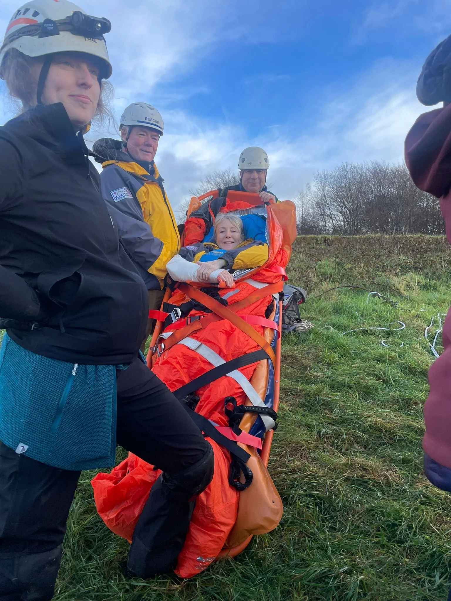 Group of rescue team members with a patient on a stretcher outdoors in a grassy area, wearing helmets and rescue gear.