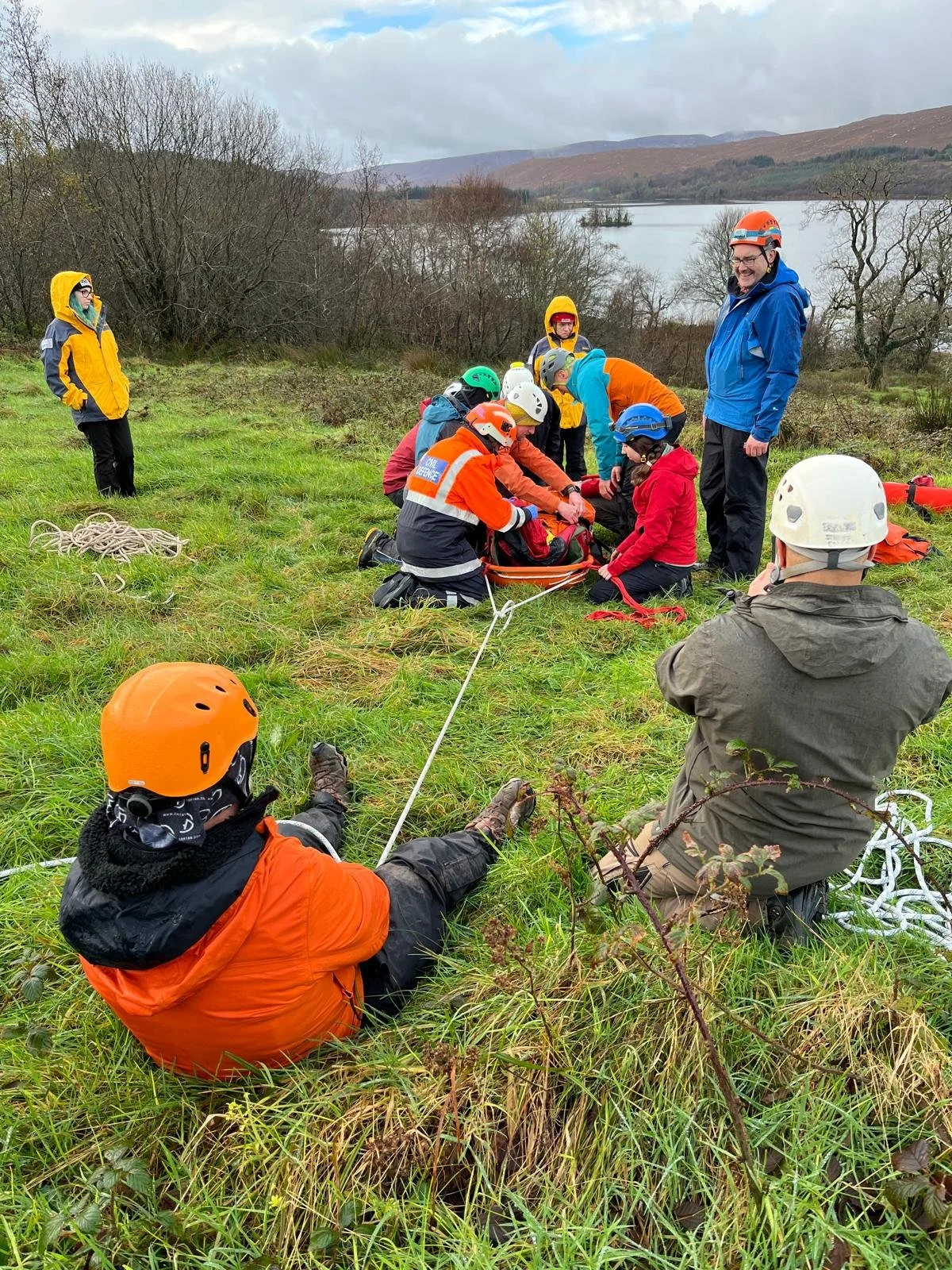 A group of people, some wearing helmets and outdoor gear, gathered around someone lying on the ground as part of a rescue or training exercise in a grassy field near a lake.