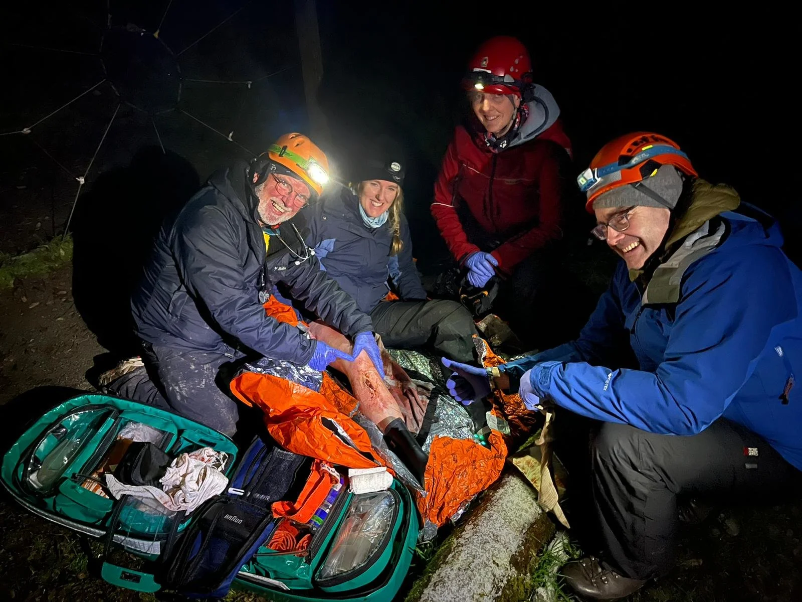 Group of four rescue team members in outdoor gear surrounding a person on a stretcher at night, attending to an injury, with rescue equipment nearby.