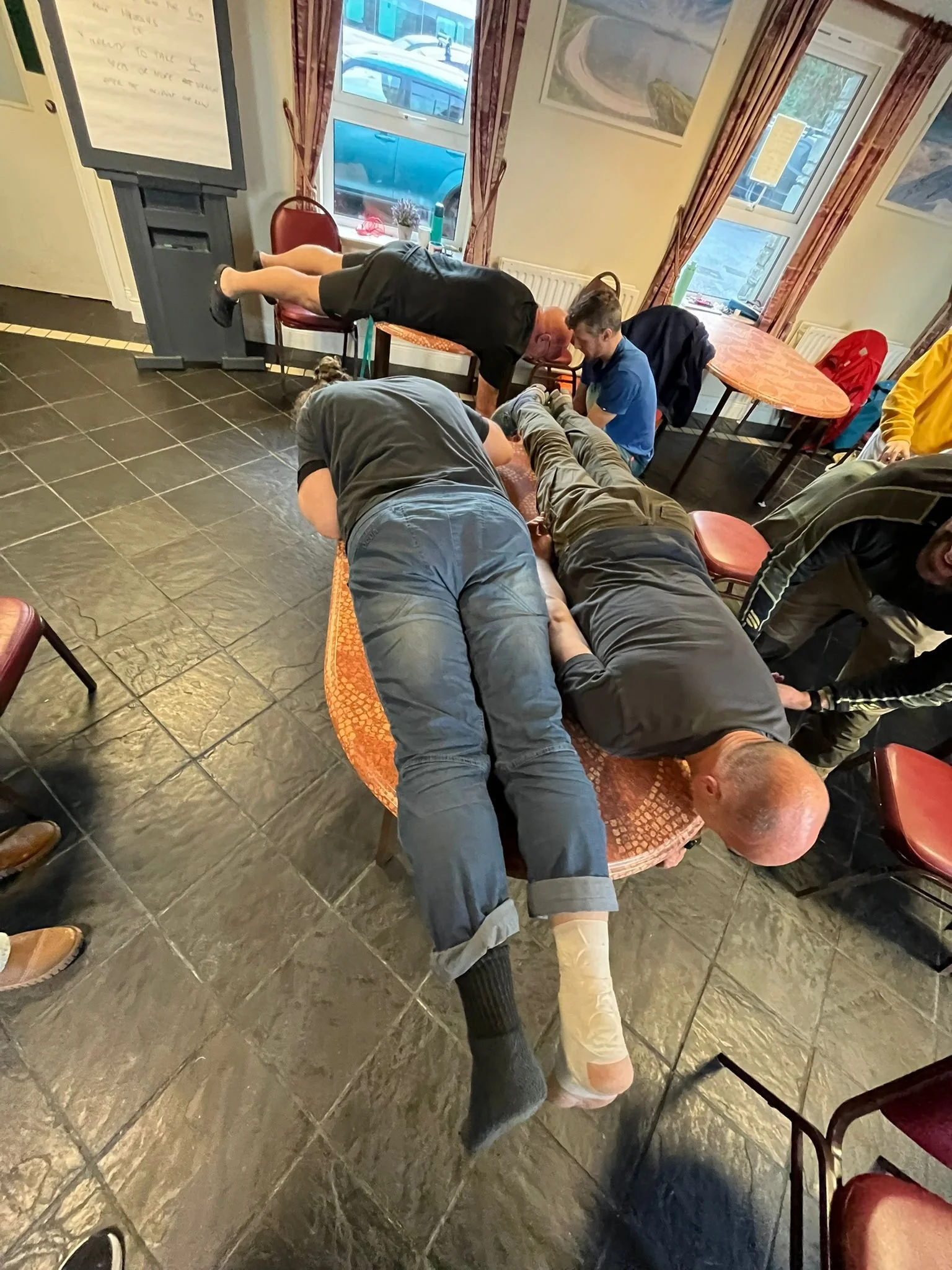 People participating in a CPR training session, practicing on a mannequin on a table in a room with large windows, curtains, and framed pictures.