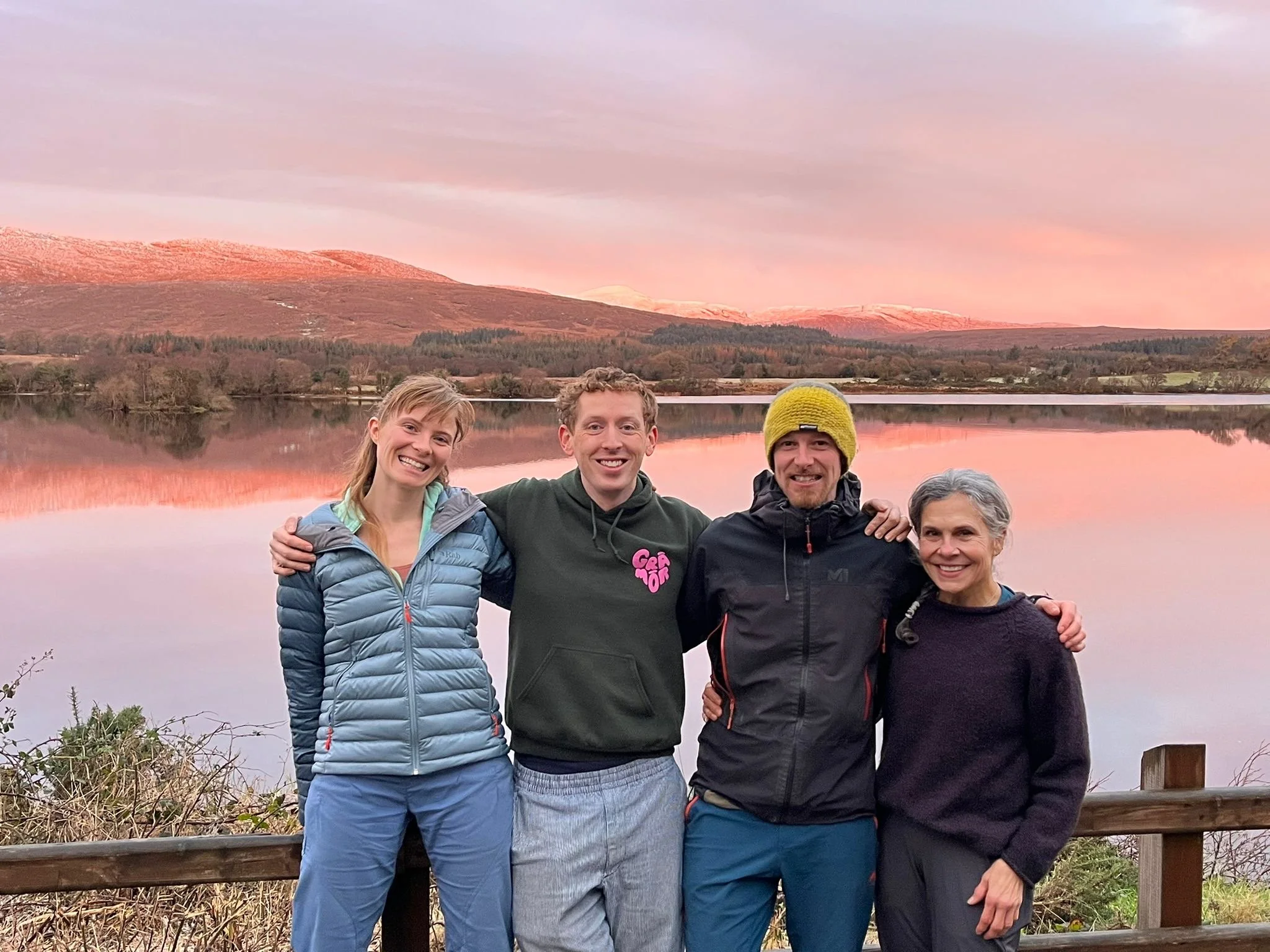 Four smiling people standing outdoors by a lake at sunset, with mountains in the background, arms around each other.