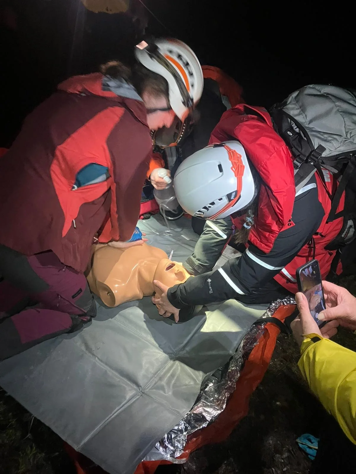 Rescue team performing CPR on a dummy during a nighttime training exercise outside, with rescue gear and backpacks around.