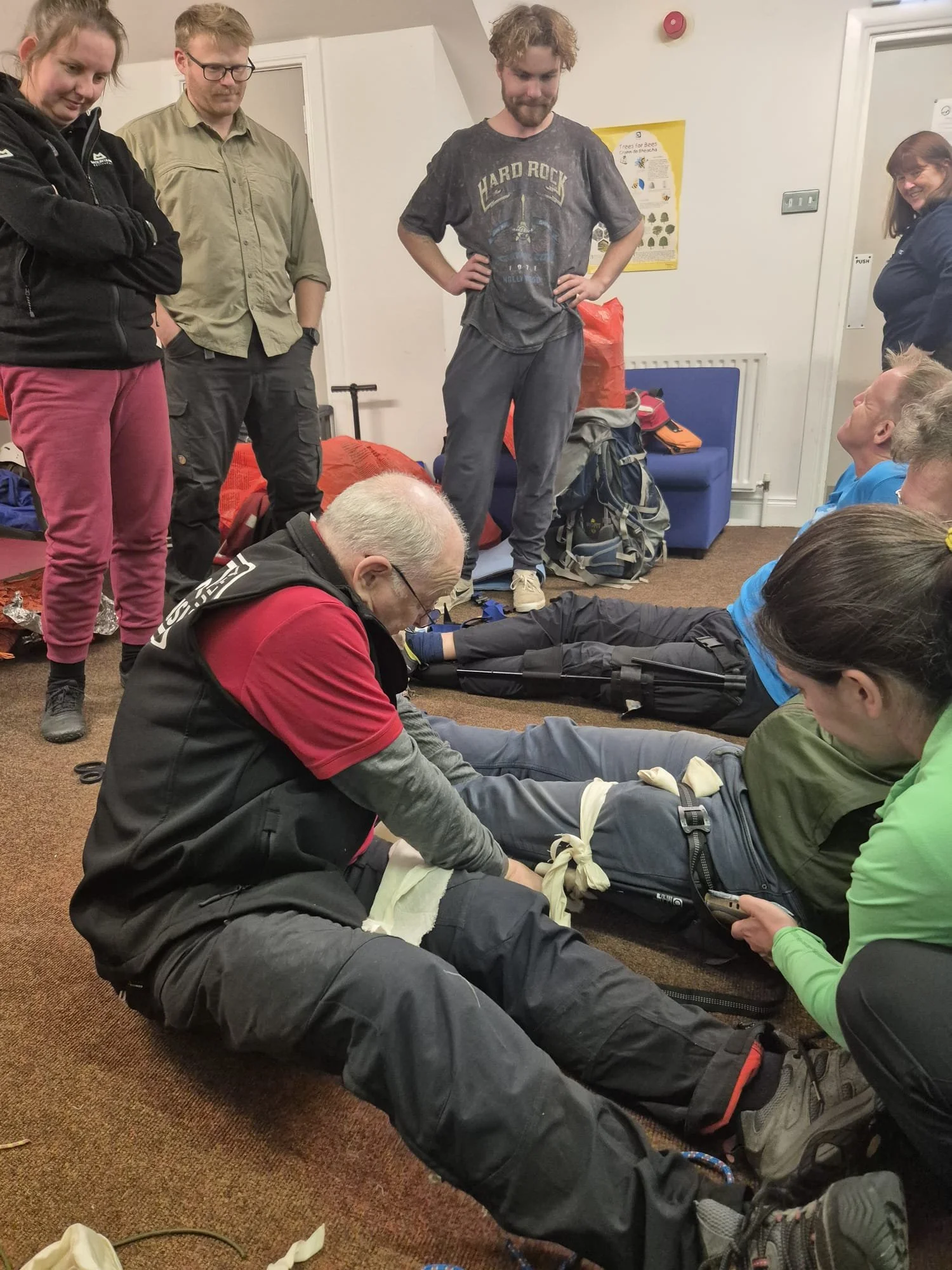 Group of people in a room, with some lying on the floor and others standing around, participating in a first aid training session. An instructor is demonstrating a medical procedure on one of the participants.