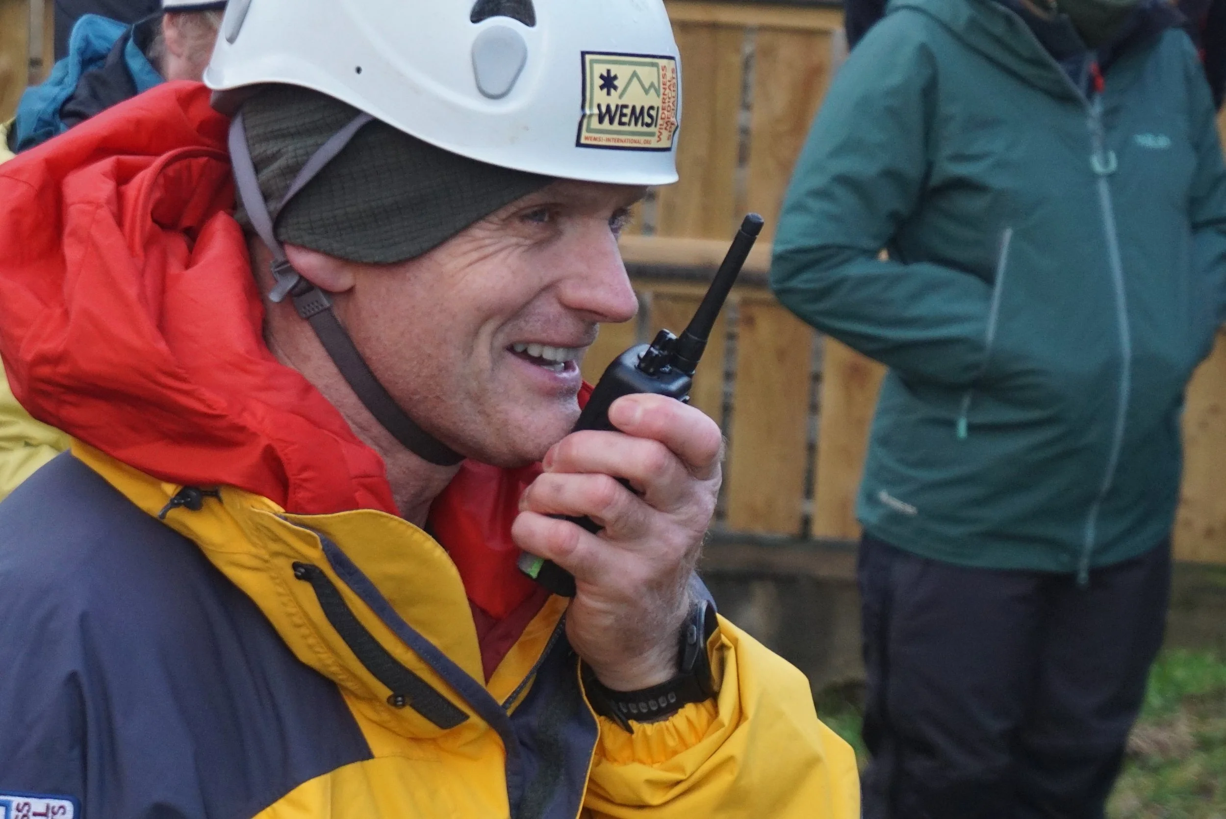 A man wearing a white helmet with a WEMSI sticker, a gray hat underneath, and a yellow and dark blue outdoor jacket is smiling while holding a walkie-talkie to his mouth. other individuals in outdoor jackets are in the background, and a wooden fence 