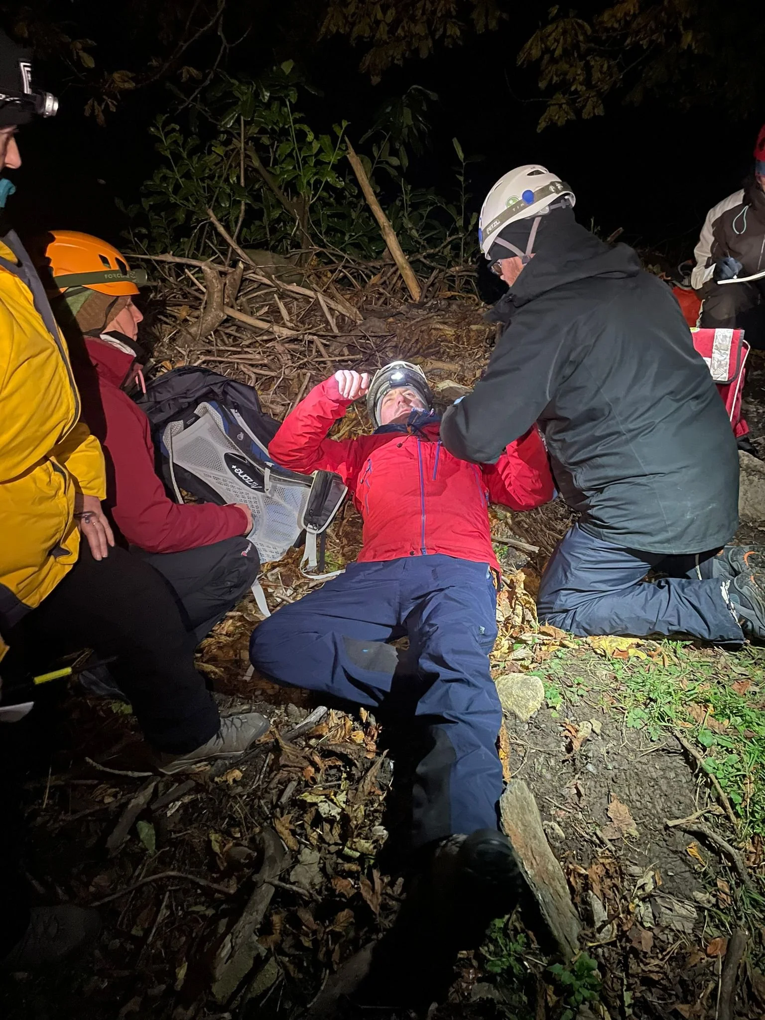 A group of rescue workers assisting an injured person lying on the ground in a forested area at night, with headlamps and safety gear.