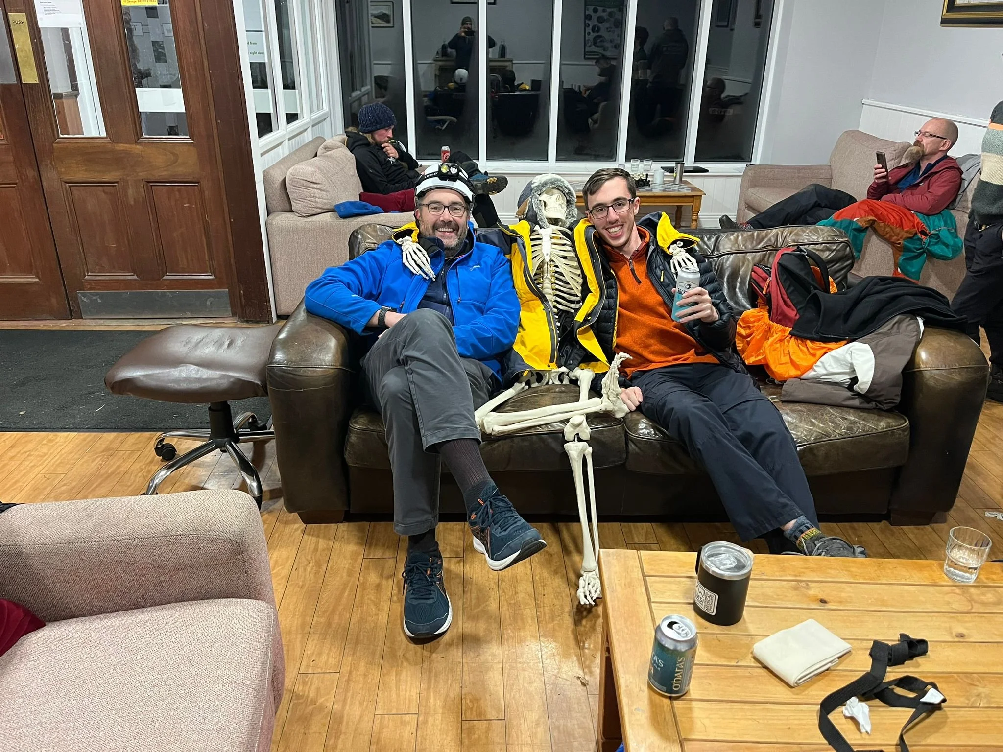 Two men sitting on a dark brown leather couch with a skeleton between them, smiling at the camera inside a cozy room with wooden floors, other people in the background, and a wooden coffee table with drinks and belongings.